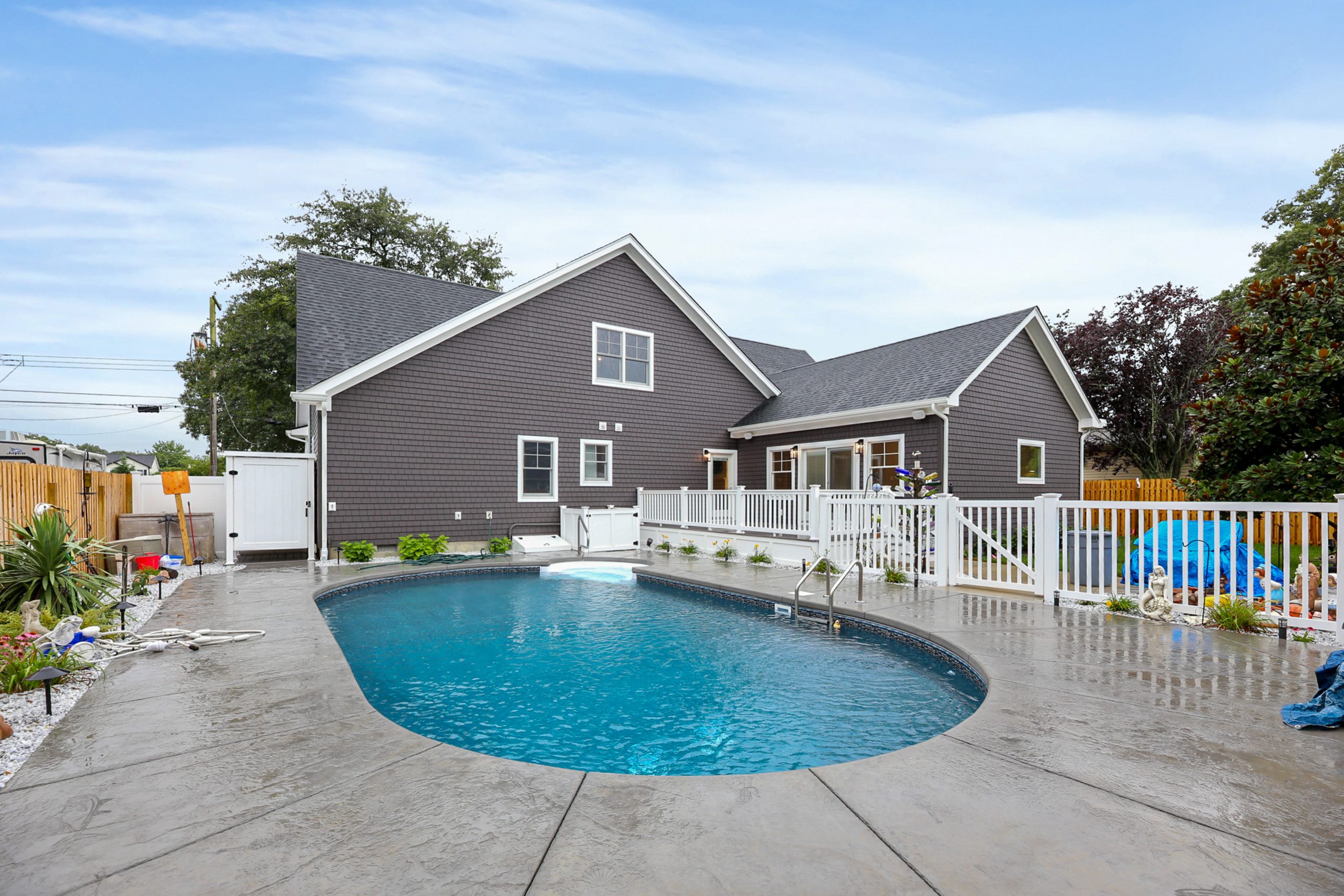 A modern house with gray siding and white trim sits behind a fenced, in-ground backyard pool surrounded by a concrete patio. The area is landscaped with plants and appears clean and inviting under a blue sky.