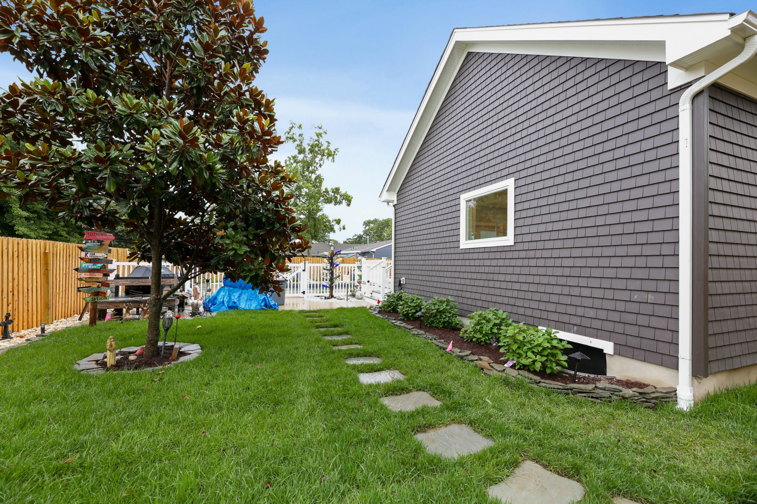 A side yard with green grass, stone pavers, a large tree, and garden beds lines the brown shingle siding of a house. A wooden fence and white gate are visible in the background.