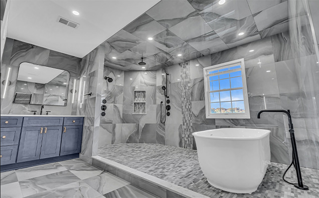 Modern bathroom with gray marble walls and floors, double vanity with blue cabinets and twin sinks, spacious walk-in shower with dual showerheads, and a white freestanding bathtub near a large window.
