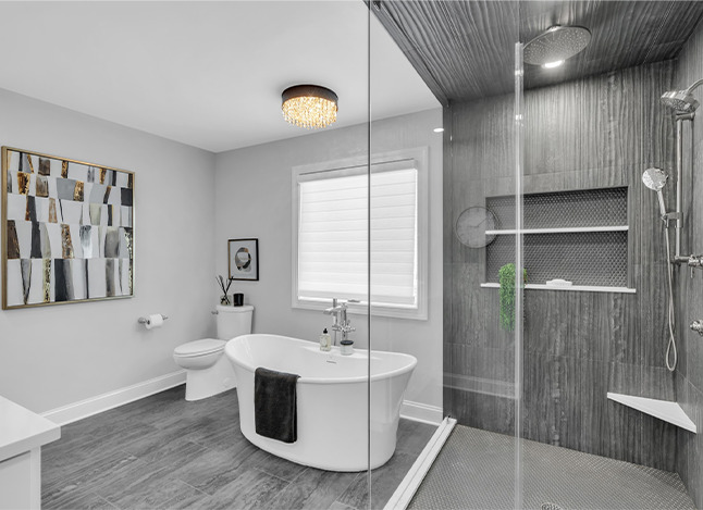 Modern bathroom with a freestanding bathtub, glass-enclosed shower, gray tile walls and floor, wall art, a toilet, and a window with a white shade. A black towel hangs on the tub and a plant decorates the shower shelf.