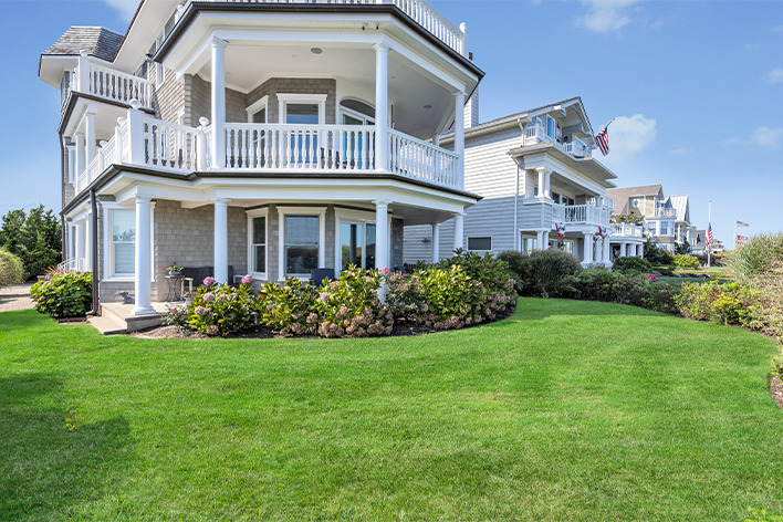 Two large, multi-story beachfront houses with wraparound balconies and manicured lawns. The sky is clear and blue, and American flags are displayed on some of the homes. Shrubs and flowers line the gardens.