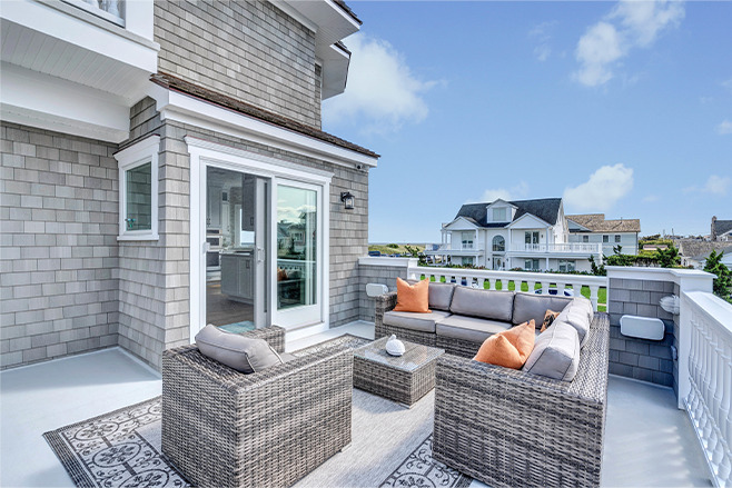 A spacious outdoor patio with gray wicker furniture, orange and gray cushions, a small table, and a patterned rug. The area overlooks neighboring houses and greenery under a bright, blue sky.