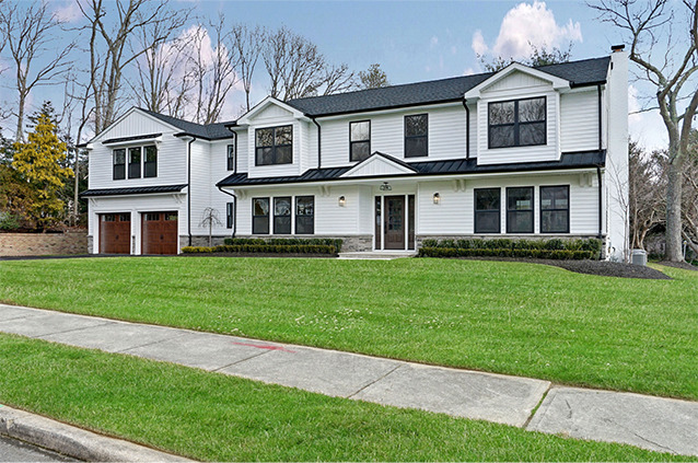 Large two-story white house with black trim, oversized windows, and a double garage with wooden doors. The home sits on a spacious, well-kept lawn along a suburban street, with trees in the background.