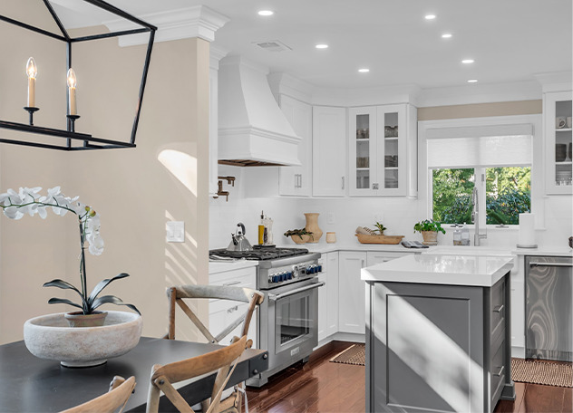 Bright, modern kitchen with white cabinets, a gray island, stainless steel appliances, and wood flooring. A dining area with a round table and chairs is in the foreground. Natural light streams in through a window.