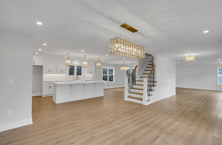 Spacious modern open-plan interior with light wood flooring, white walls, a kitchen with white cabinets and pendant lights, an island, and a staircase with wooden steps and black railings. Elegant chandeliers hang from the ceiling.