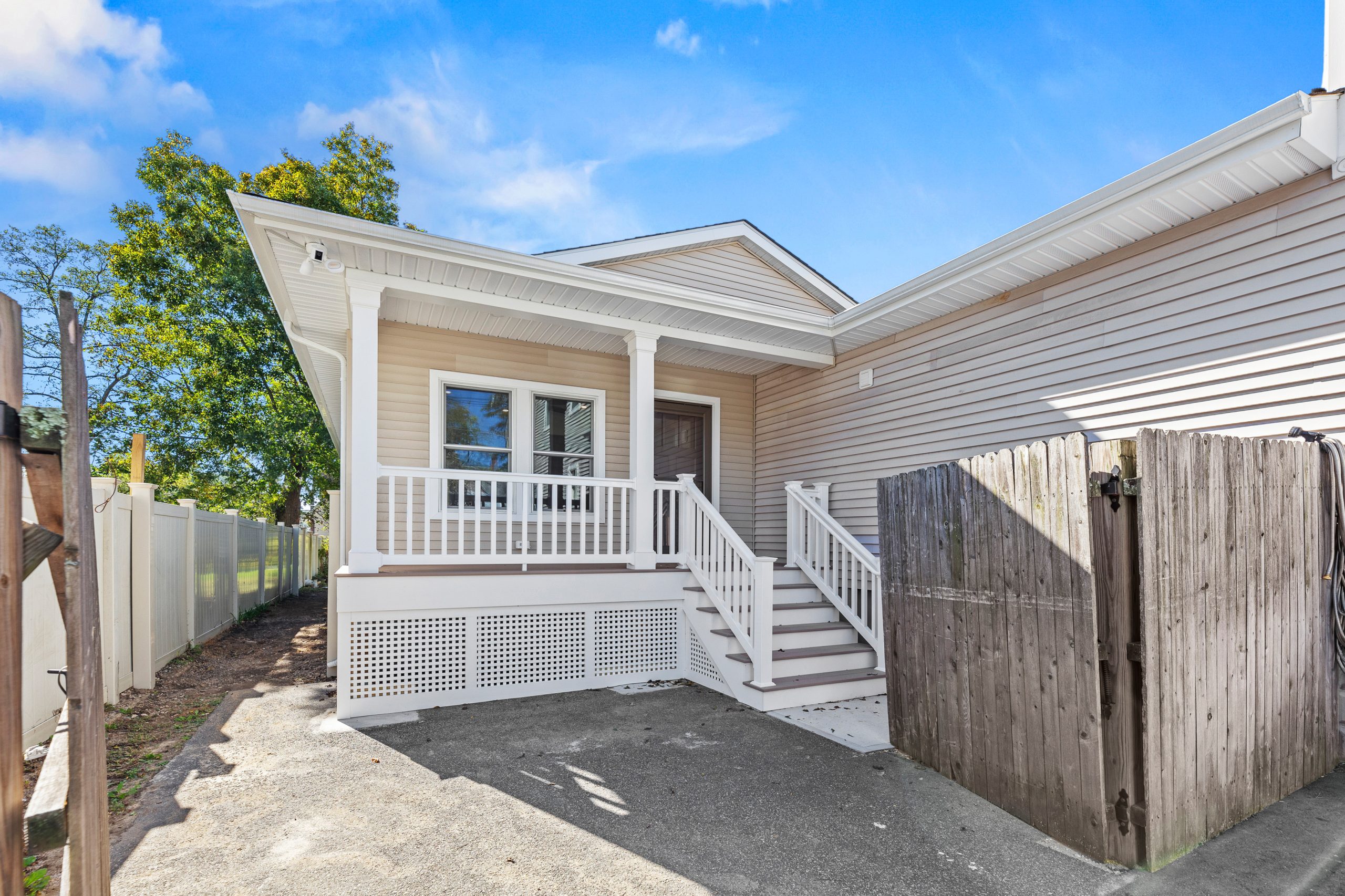 Single-story beige house with a small covered front porch, white railing, stairs leading to the front door, and a wooden gate on the right. The house sits beside a paved driveway and a white fence.