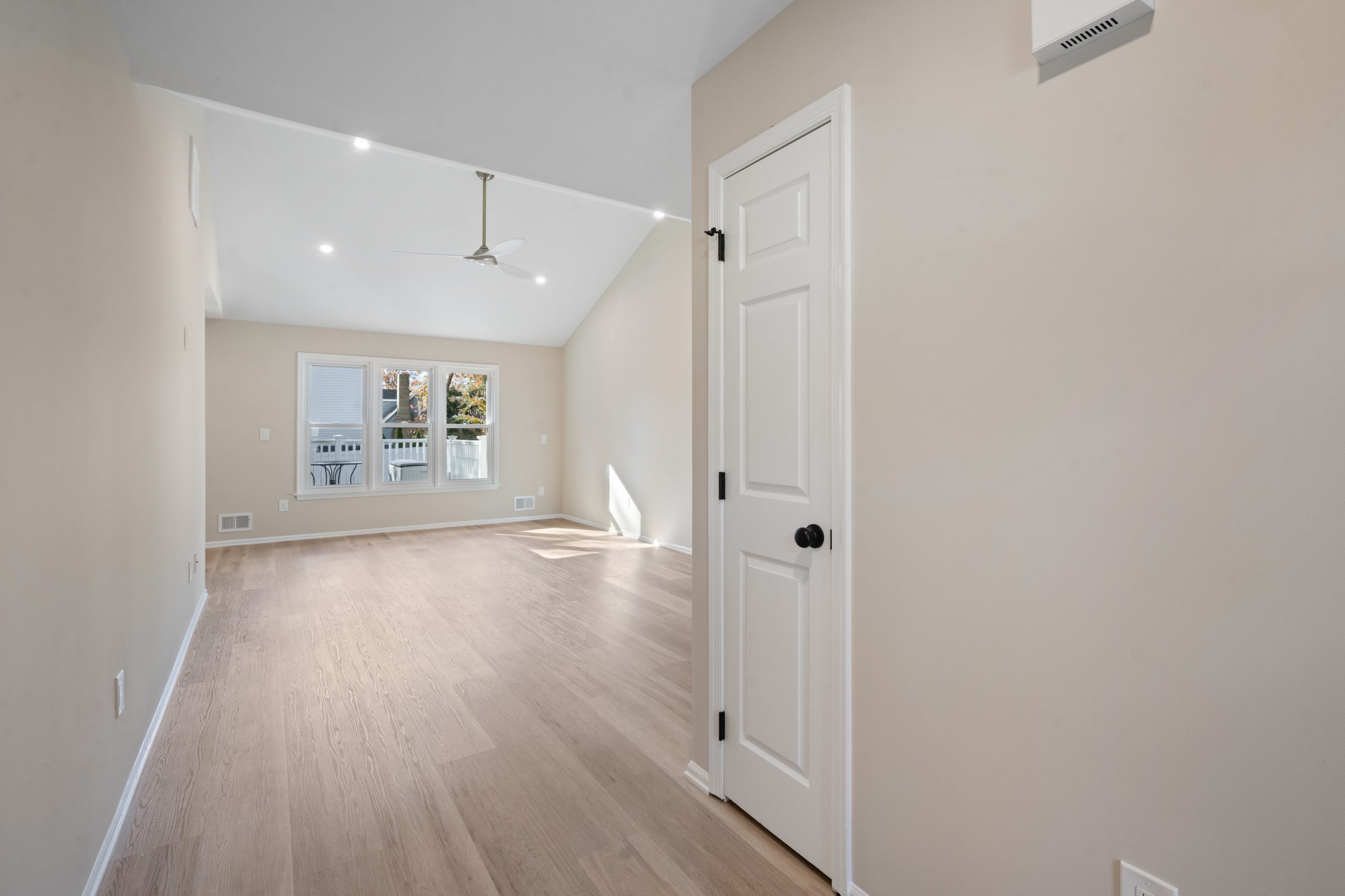 Spacious, empty room with light wood floors, beige walls, white ceiling, and a ceiling fan. A white door is on the right, and large windows at the far end let in natural light.