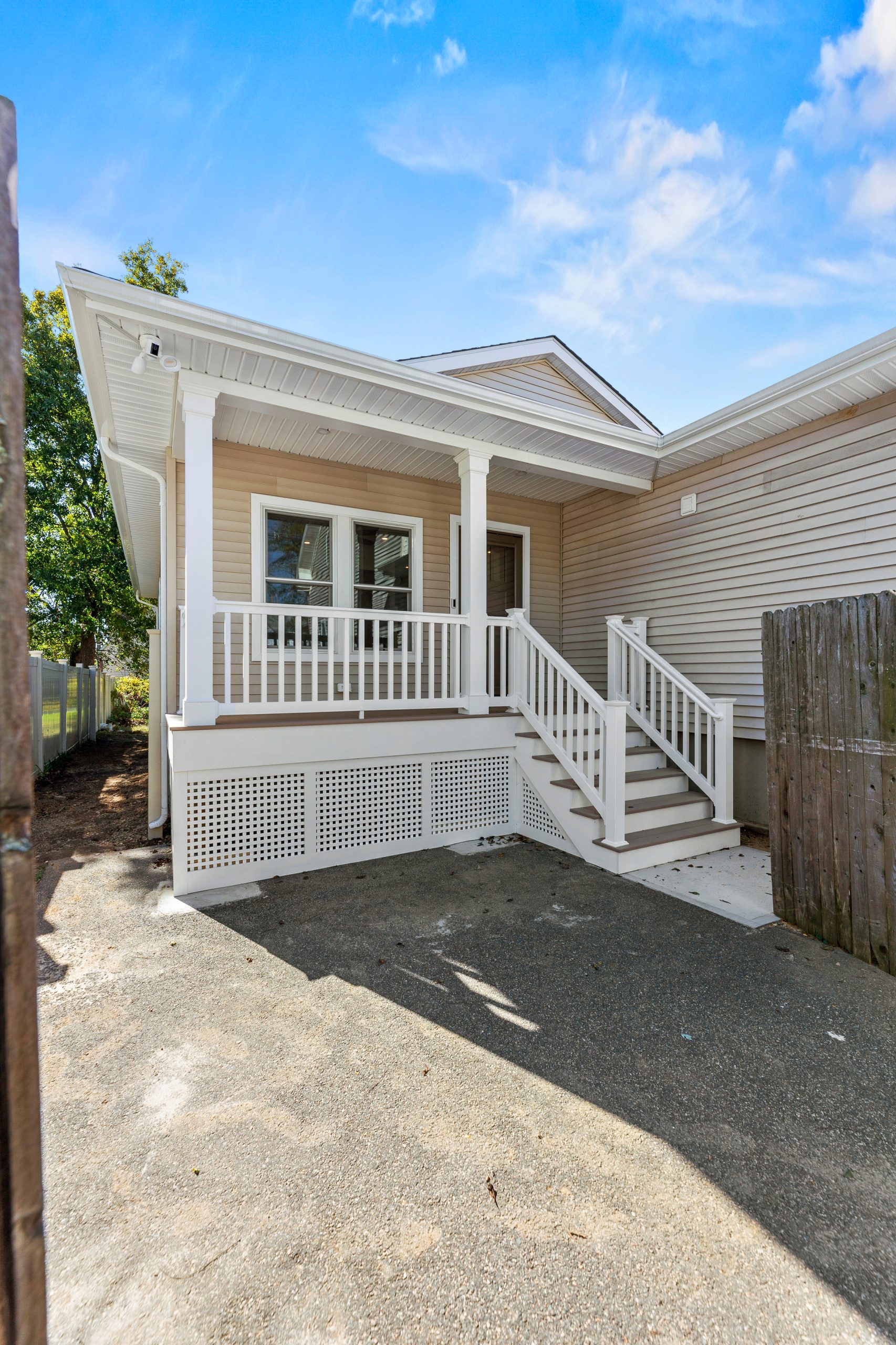 A beige house with white trim features a covered porch, white railings, and steps leading down to a gravel driveway. There is a wooden fence on the right and trees in the background under a blue sky with clouds.