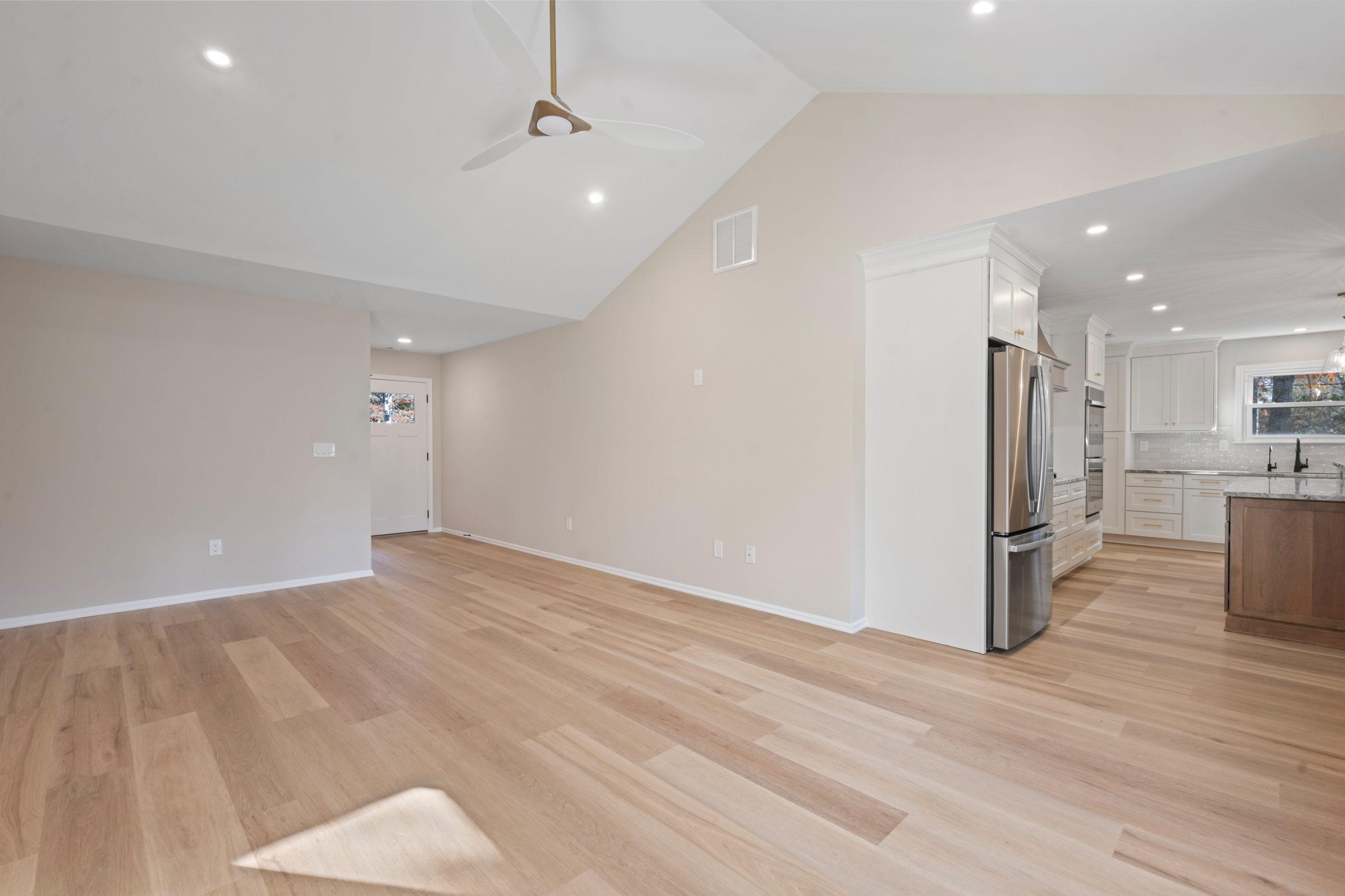 Open-concept living area with light wood flooring, vaulted white ceiling, ceiling fan, and recessed lights. A modern kitchen with stainless steel appliances is visible to the right. The entry door is at the back.