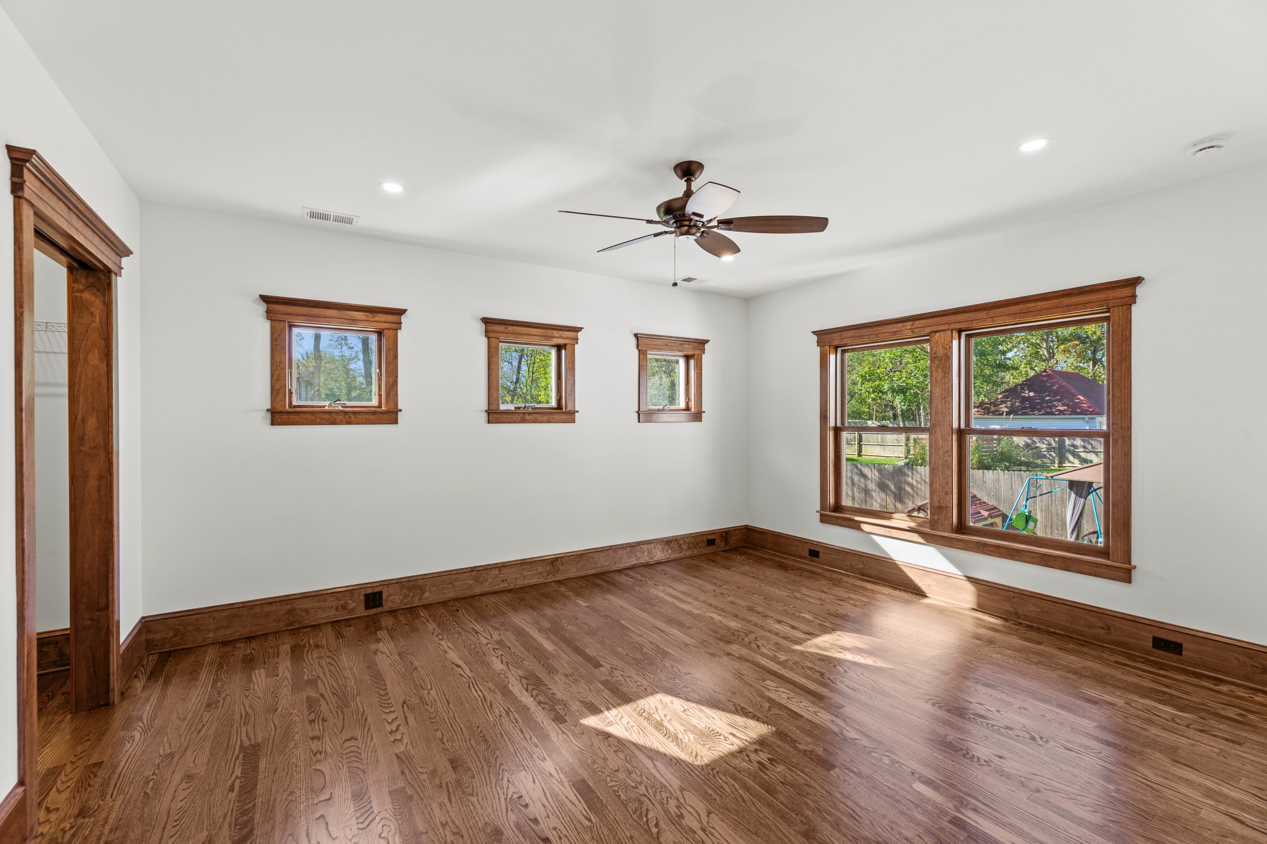 Bright, empty room with white walls, wooden trim, four small rectangular windows on one wall, two larger windows on another, hardwood floors, and a ceiling fan with lights. Trees and a yard visible through the windows.