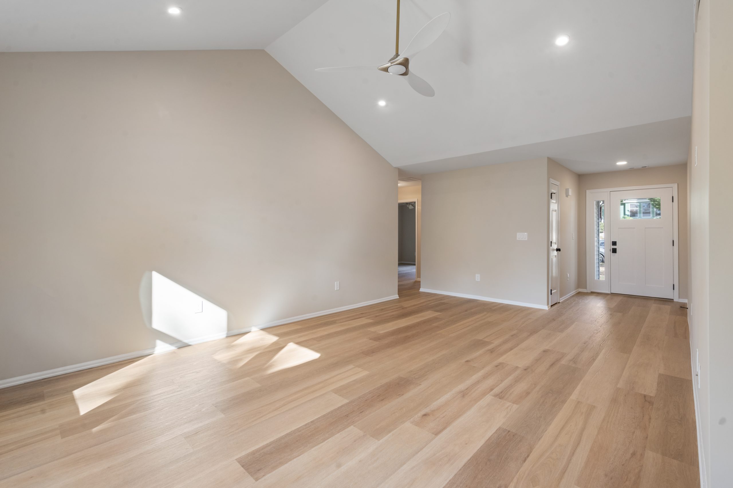 A bright, empty living room with light wood flooring, beige walls, a vaulted ceiling with a white ceiling fan, recessed lighting, and natural light streaming in from a window near the front door.