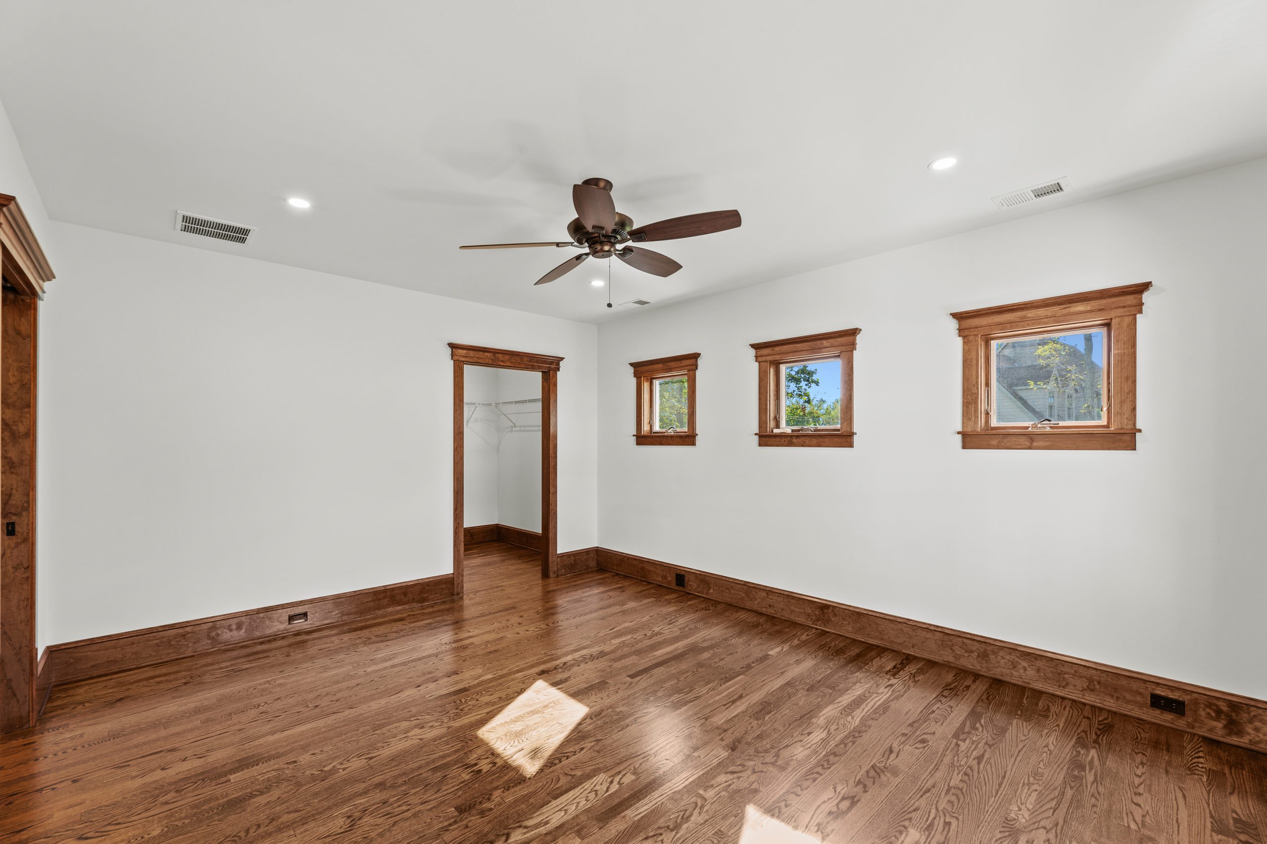 Empty bedroom with hardwood floors, white walls, three rectangular windows with wood trim, a ceiling fan with lights, and an open doorway leading to a walk-in closet.