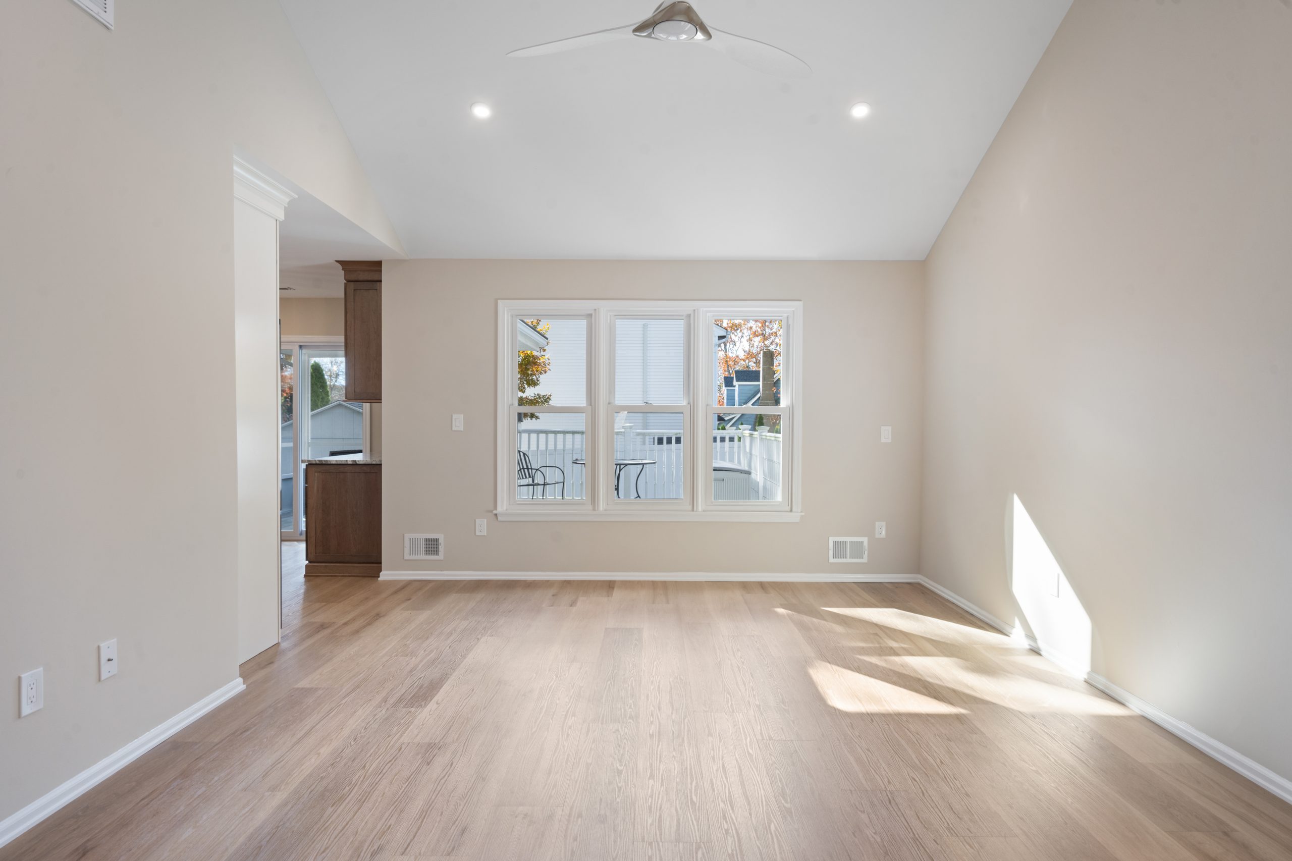 Bright, empty living room with light wood floors, beige walls, vaulted ceiling with a ceiling fan, and three large windows letting in natural light; kitchen visible through an open doorway on the left.
