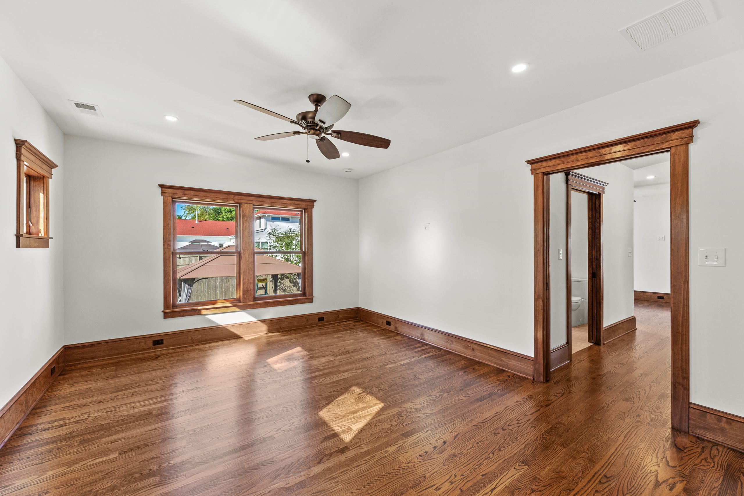 Bright, empty room with hardwood floors, white walls, wood trim, a ceiling fan, large window, and an open doorway leading to a hallway. Sunlight enters through the window, illuminating the space.