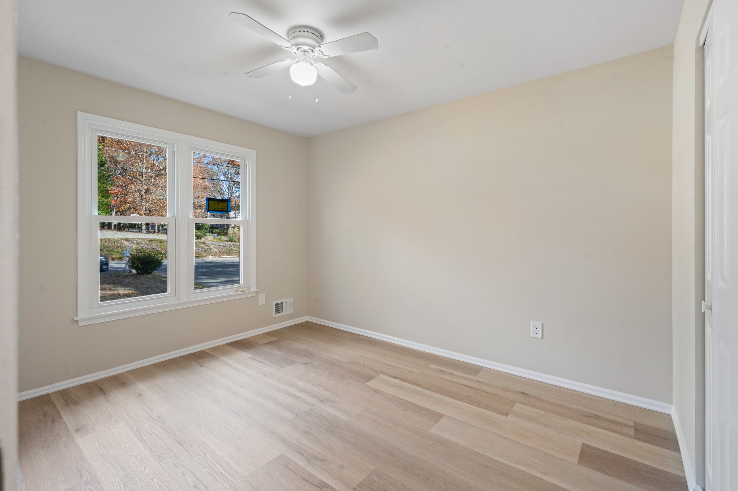 A small, empty room with light wood flooring, beige walls, a white ceiling fan, a double window letting in natural light, and a white electrical outlet on the wall. Trees with autumn leaves are visible outside.