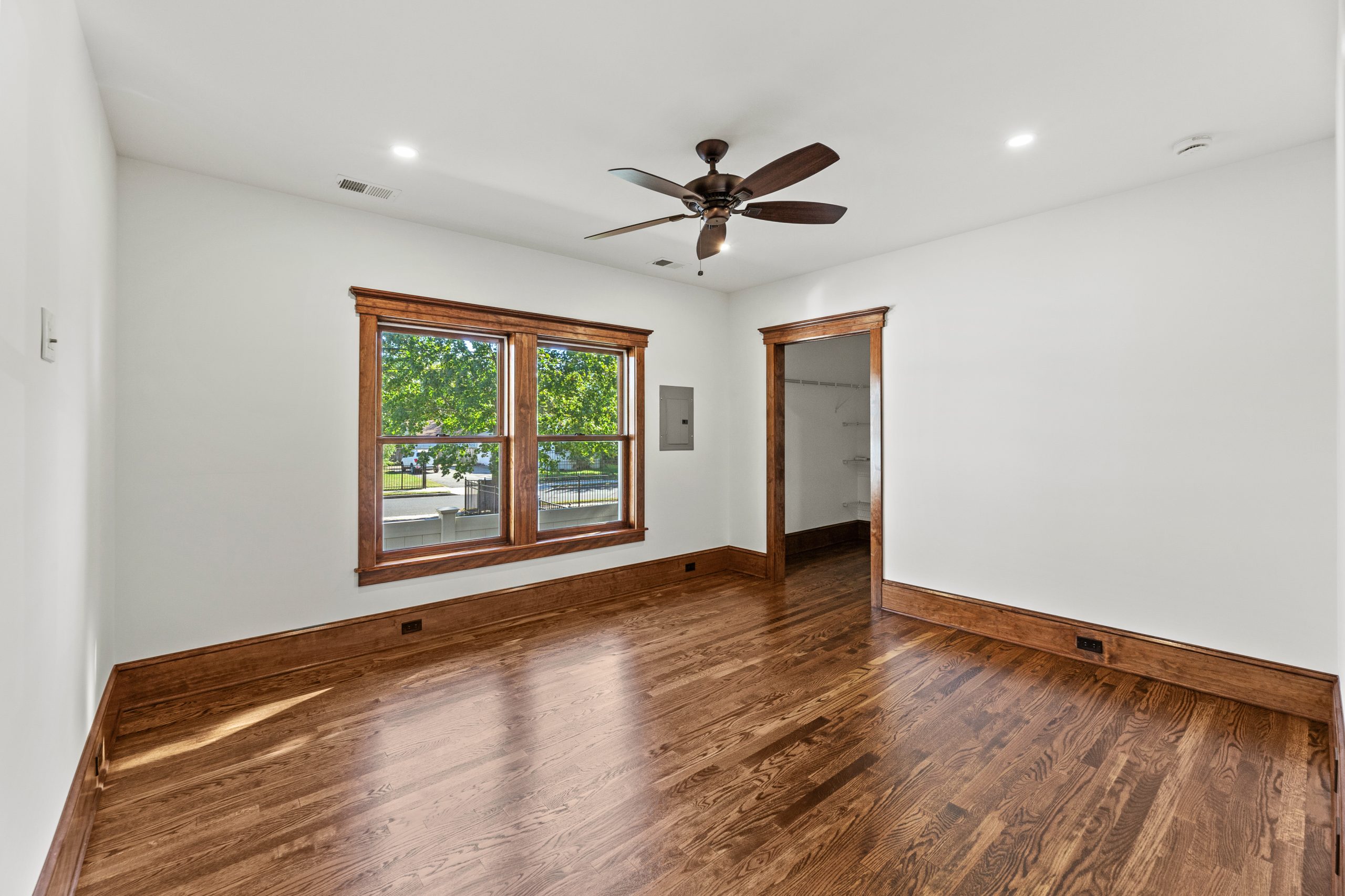A bright, empty room with hardwood floors, white walls, a ceiling fan, large window with wooden trim, and a wooden doorframe leading to another space. Sunlight streams in through the window.