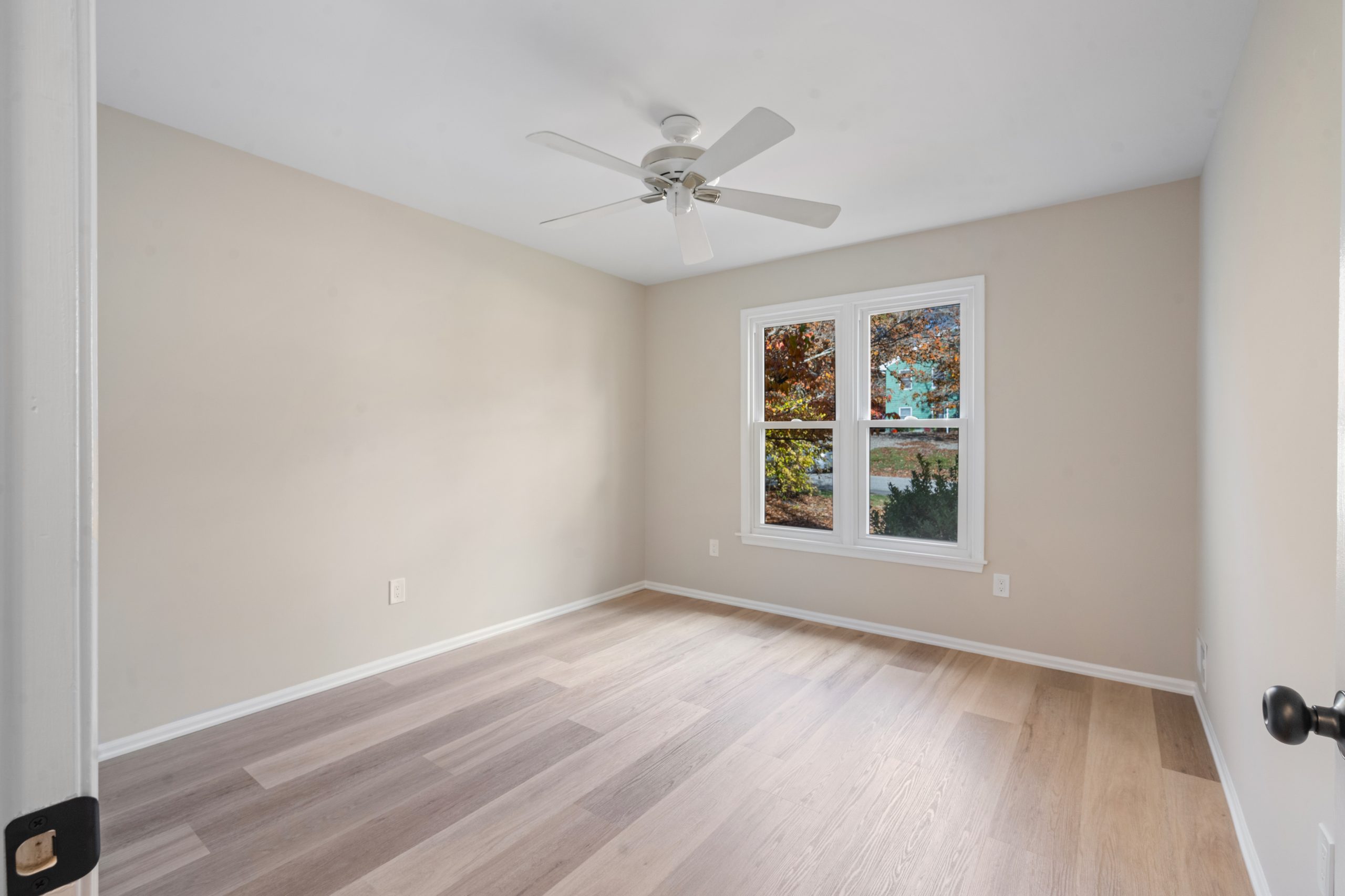 A bright, empty room with light wood flooring, beige walls, a white ceiling fan, and a window showing trees and fall foliage outside.
