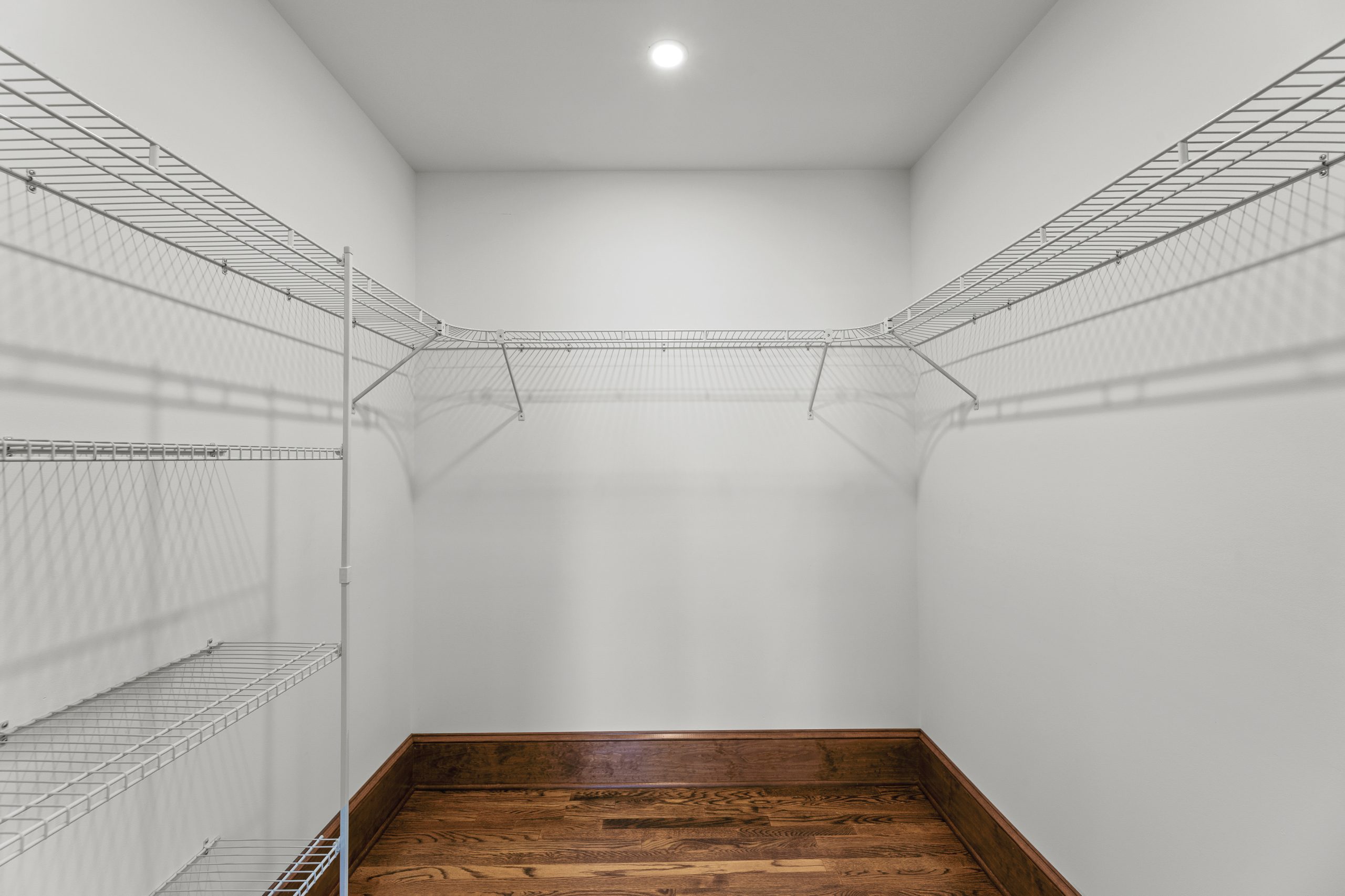 A spacious, empty walk-in closet with white wire shelving on three sides, white walls, and hardwood floors. A ceiling light illuminates the space.