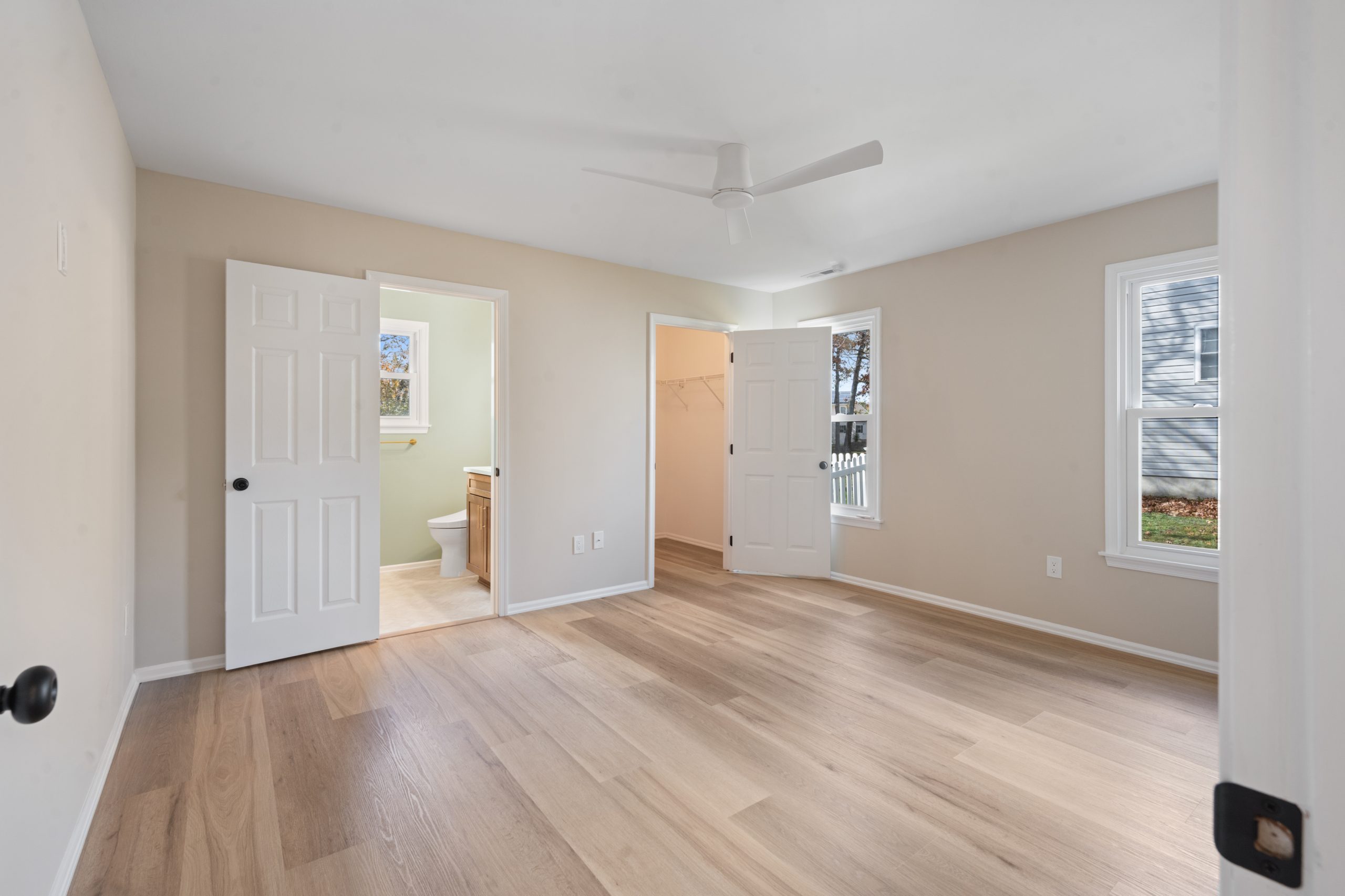 Spacious, empty bedroom with light wood floors, beige walls, white ceiling fan, two windows, and two open doors leading to a bathroom and a walk-in closet. Natural light fills the room.
