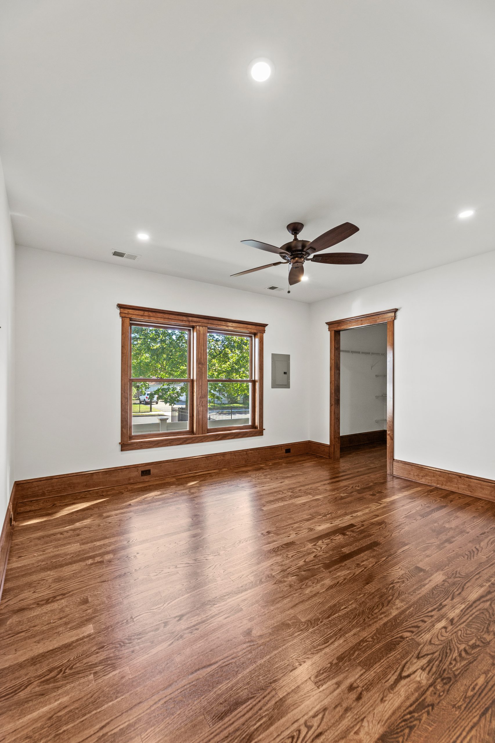 An empty room with hardwood floors, white walls, a ceiling fan, and large double windows framed in wood. Sunlight streams through the windows, highlighting the natural wood trim and floor.