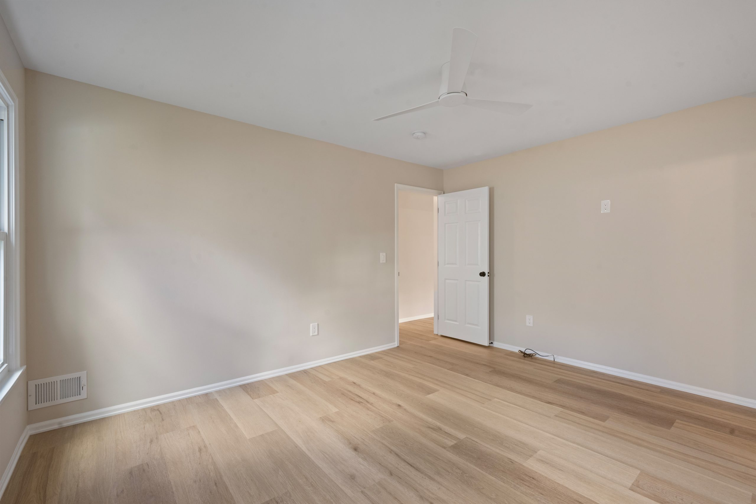 Empty room with light wood flooring, beige walls, a white ceiling fan, white baseboards, a window on the left, an open white door, and visible electrical outlets. Natural light brightens the space.