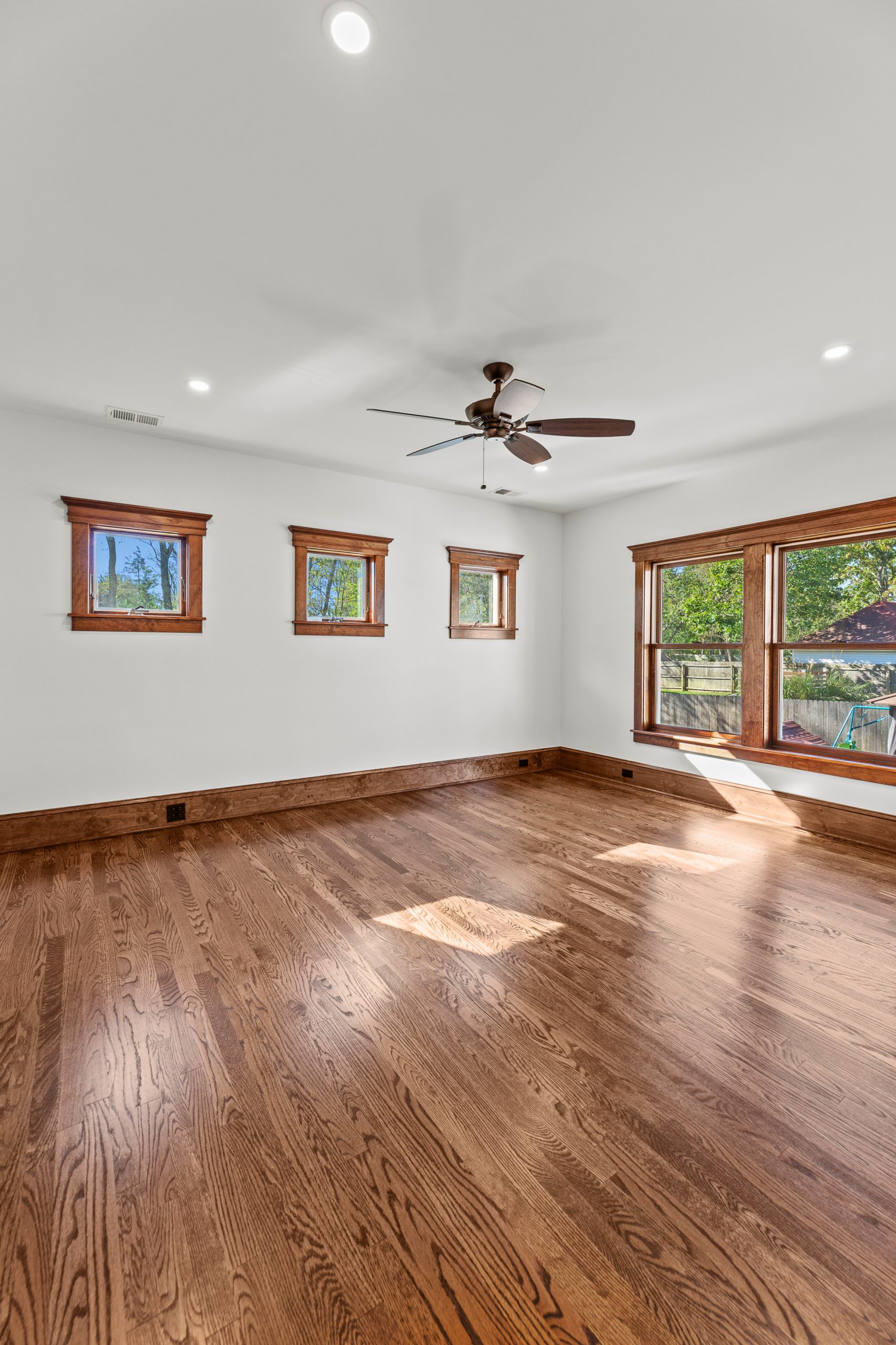 Bright empty room with polished wood floors, white walls, a ceiling fan, and wood-trimmed windows letting in natural light. Large windows overlook a green yard, and three smaller windows line one wall.