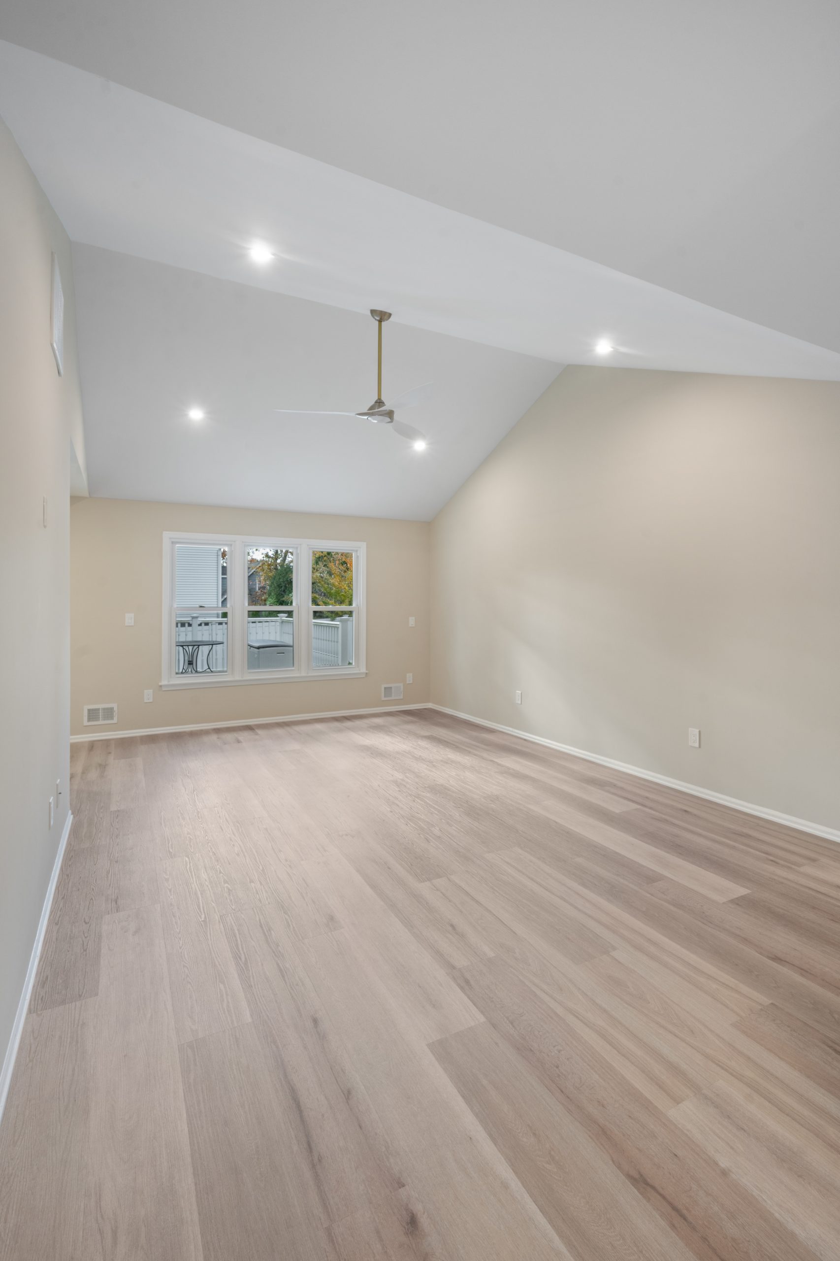 A spacious, empty room with light wood flooring, beige walls, a white ceiling, recessed lights, a ceiling fan, and three large windows leading to a balcony with a view of greenery outside.