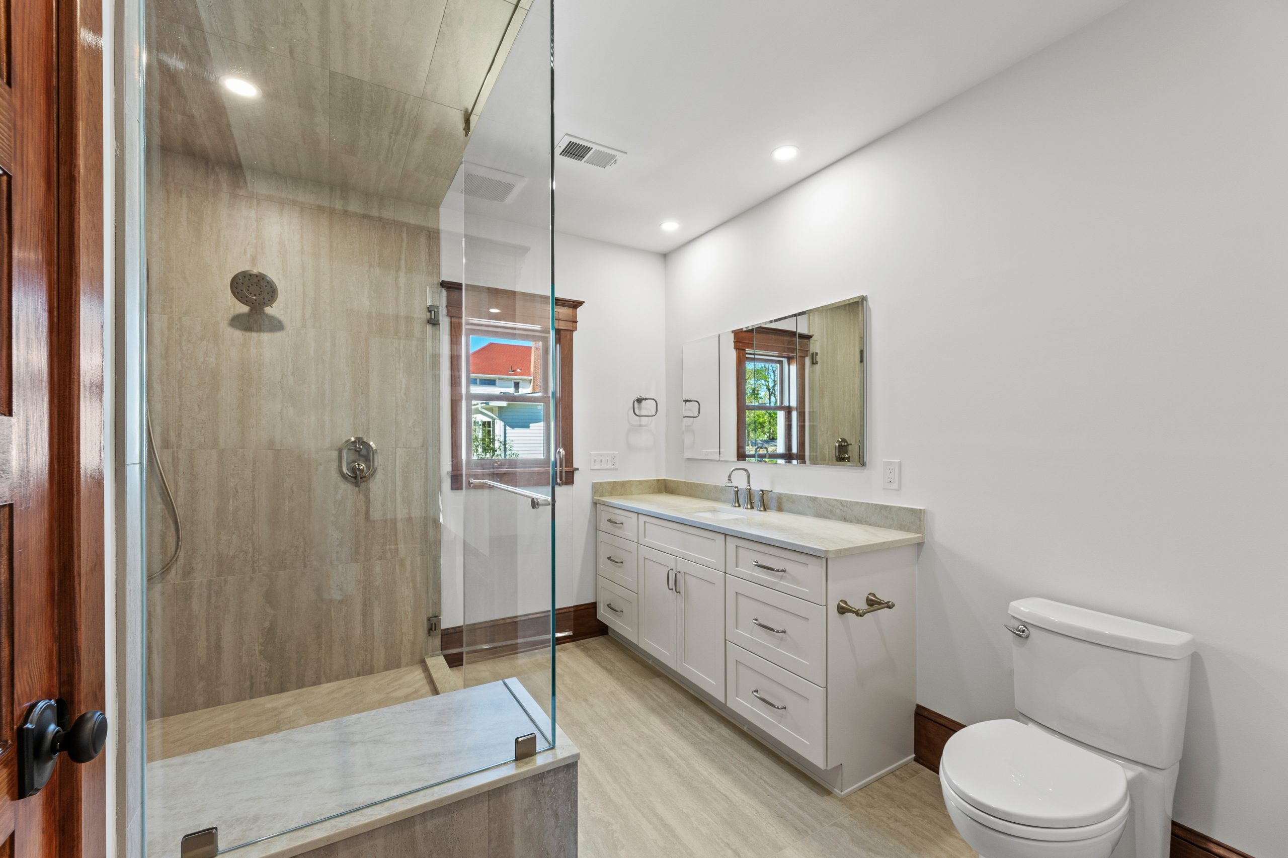 Modern bathroom with a large glass-enclosed shower, white vanity with dual sinks and a wide mirror, toilet, light walls, and natural wood accents around the windows and door.