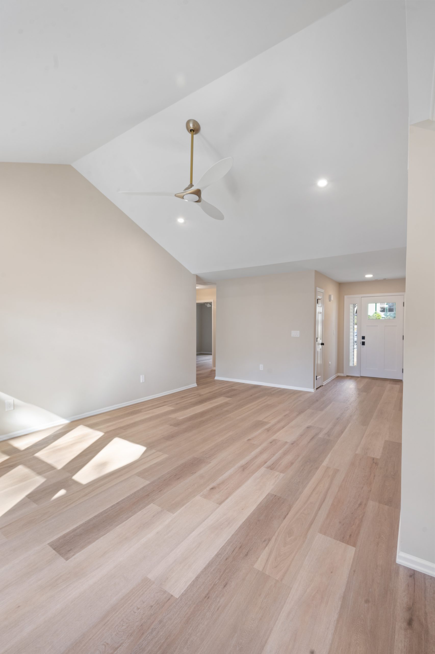 Spacious, empty room with light wood floors, vaulted white ceiling, ceiling fan, neutral-colored walls, and natural light streaming through windows near the entrance door in the background.