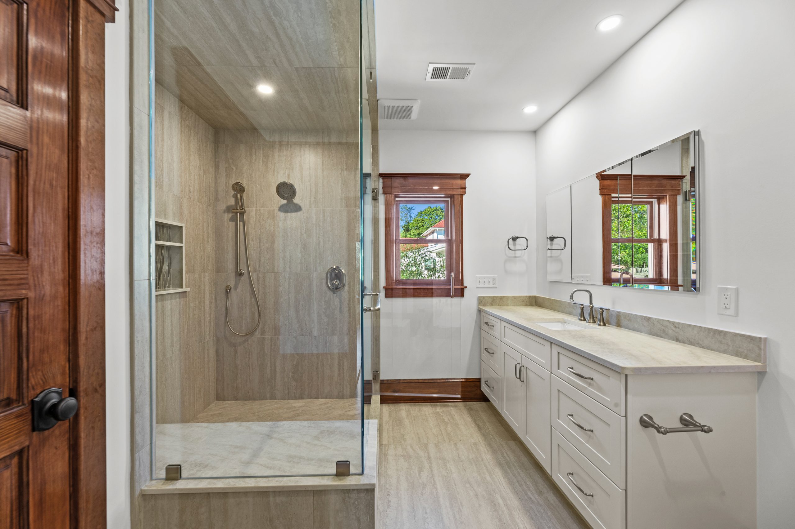 Modern bathroom with a large glass-enclosed shower, beige tile, a double-sink vanity with white cabinets, a wide mirror, and natural light from a window with wooden trim.