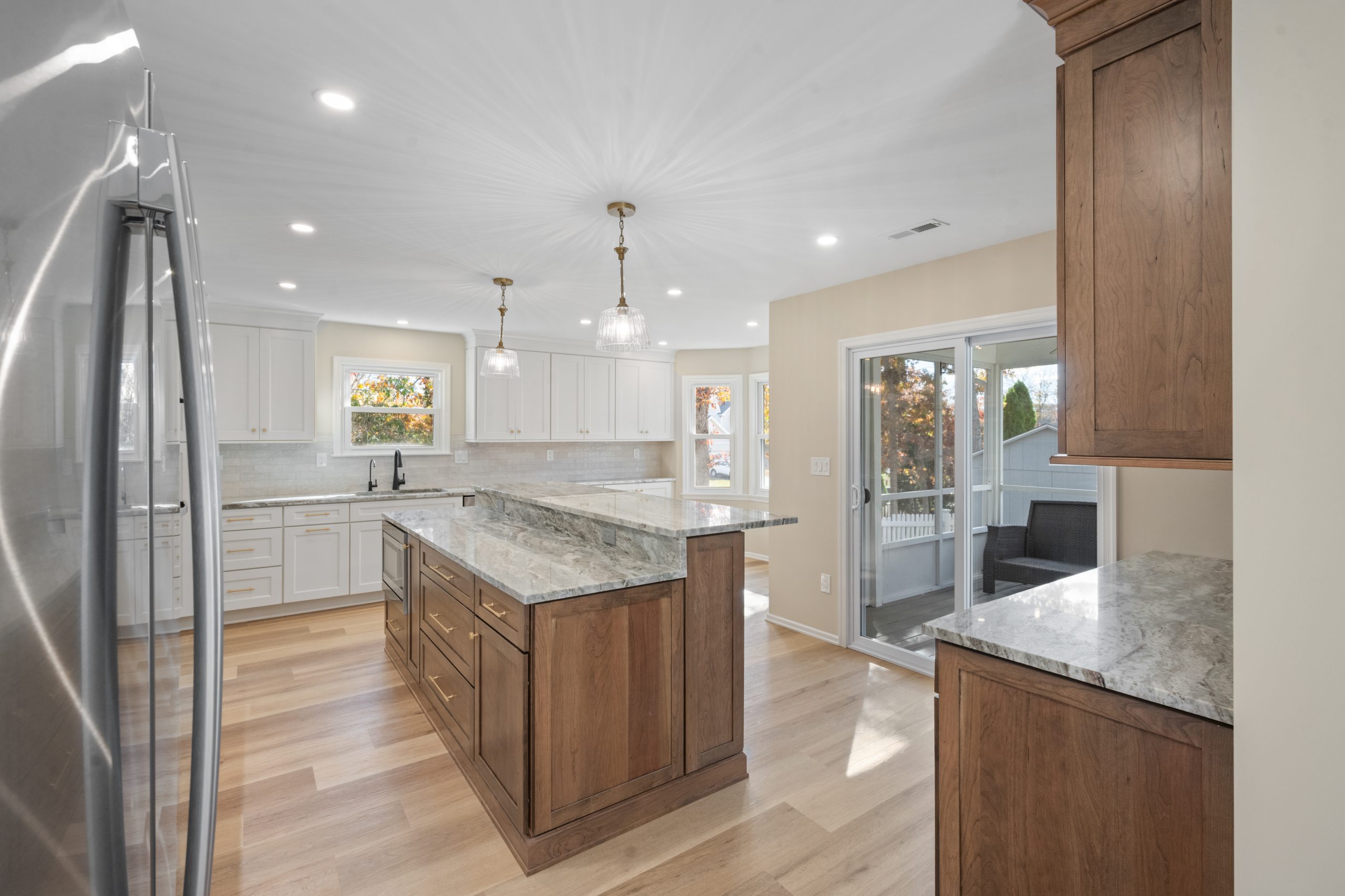 Spacious modern kitchen with wood and white cabinets, marble countertops, pendant lights, stainless steel refrigerator, island, and large sliding glass doors leading to a deck. Light wood flooring throughout.