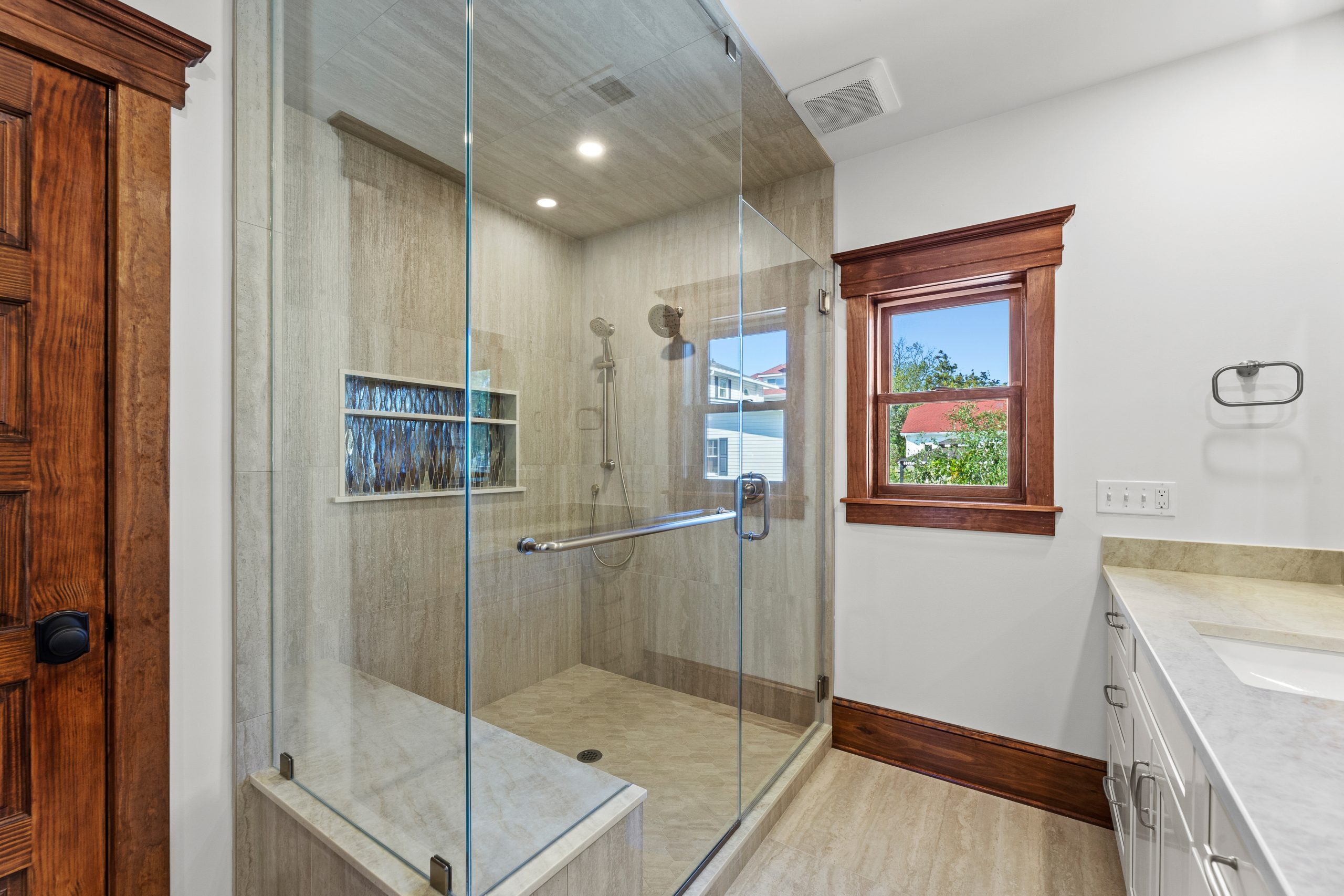 Modern bathroom featuring a spacious glass-enclosed shower with built-in shelves, beige tile walls and floor, a wooden-framed window, white countertop, and wooden accents.