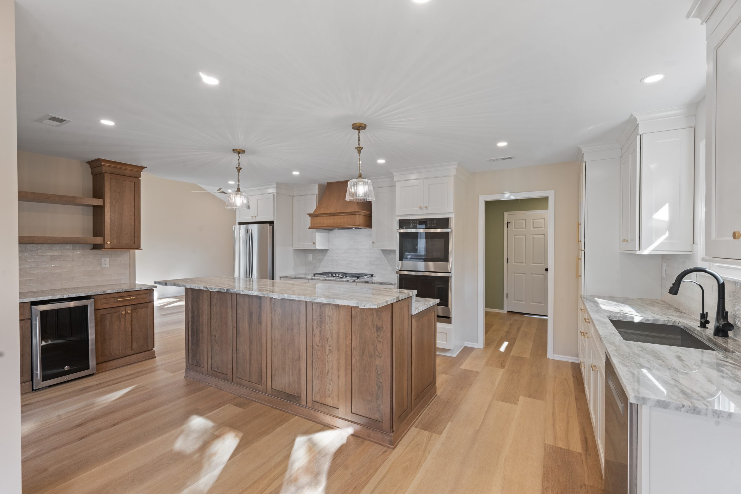 Modern kitchen with wooden floors, a large island with marble countertop, white cabinets, double ovens, stainless steel appliances, hanging pendant lights, and a sink under a window with natural light.