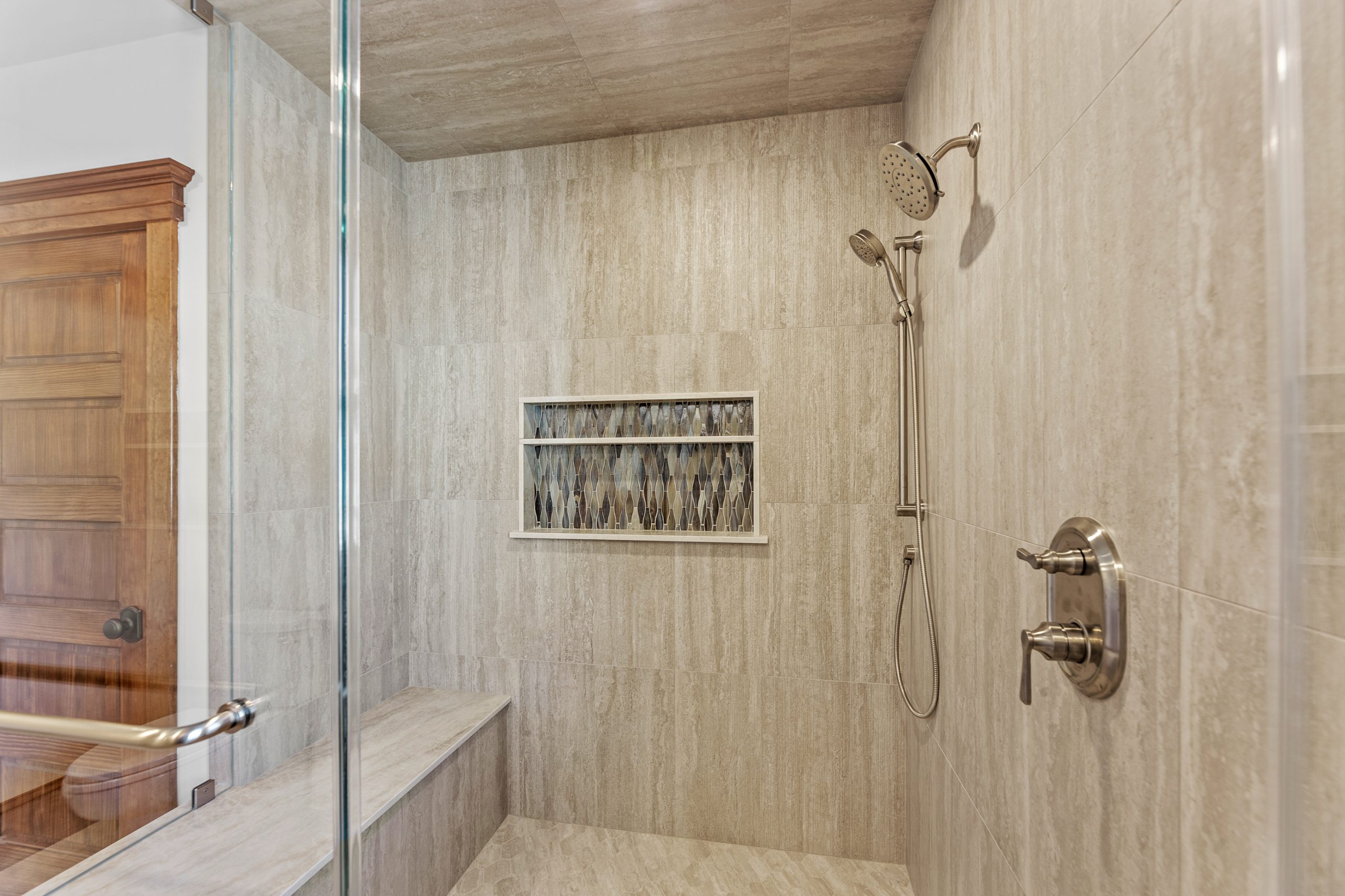 A modern walk-in shower with beige tile walls and floor, a built-in bench, a decorative niche with patterned tiles, a glass door, and brushed nickel shower fixtures. A wooden door is visible outside the shower.