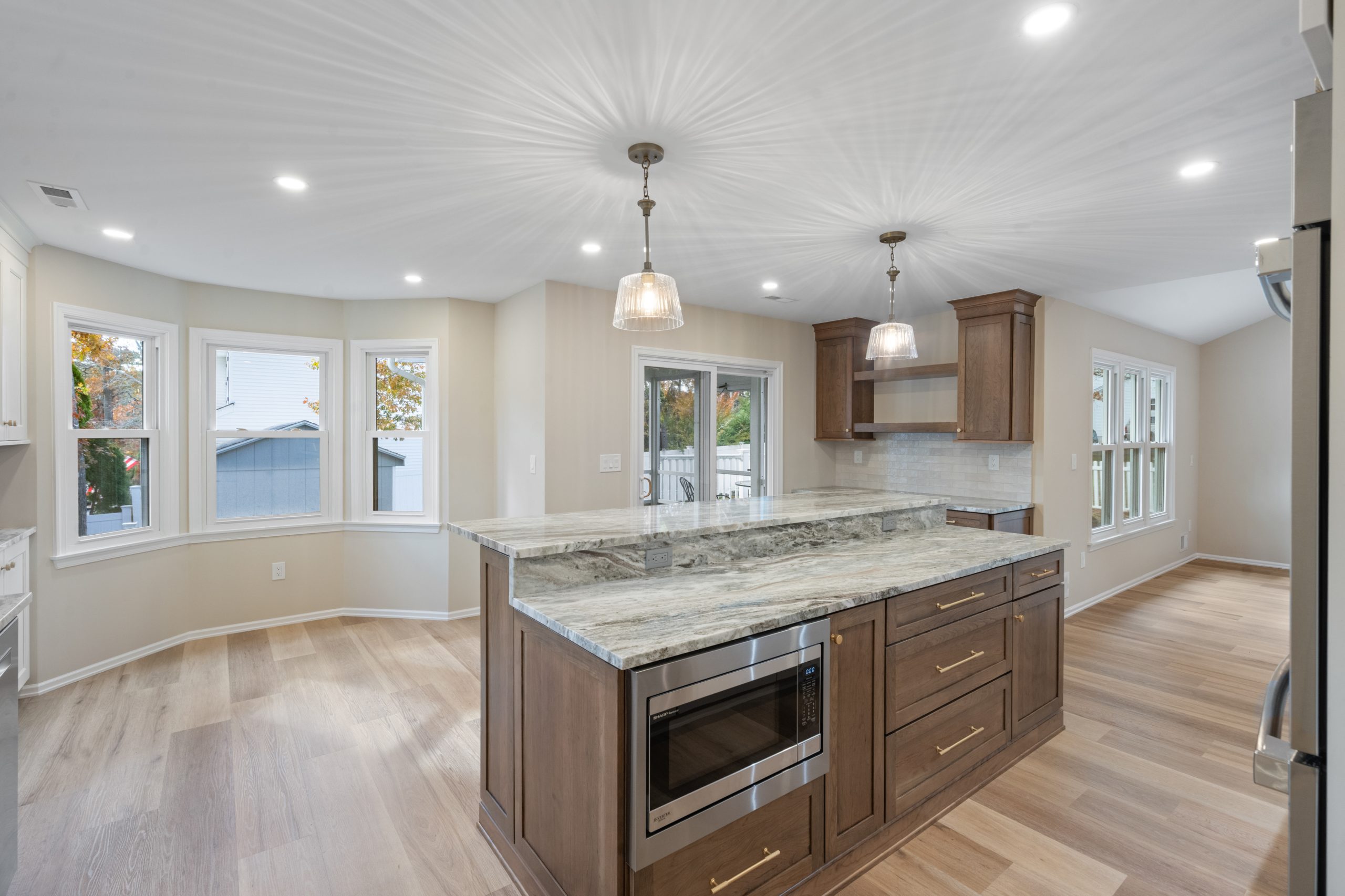 Modern kitchen with wood flooring, a large island featuring a marble countertop, built-in microwave, pendant lights, and surrounding bay windows that let in natural light.