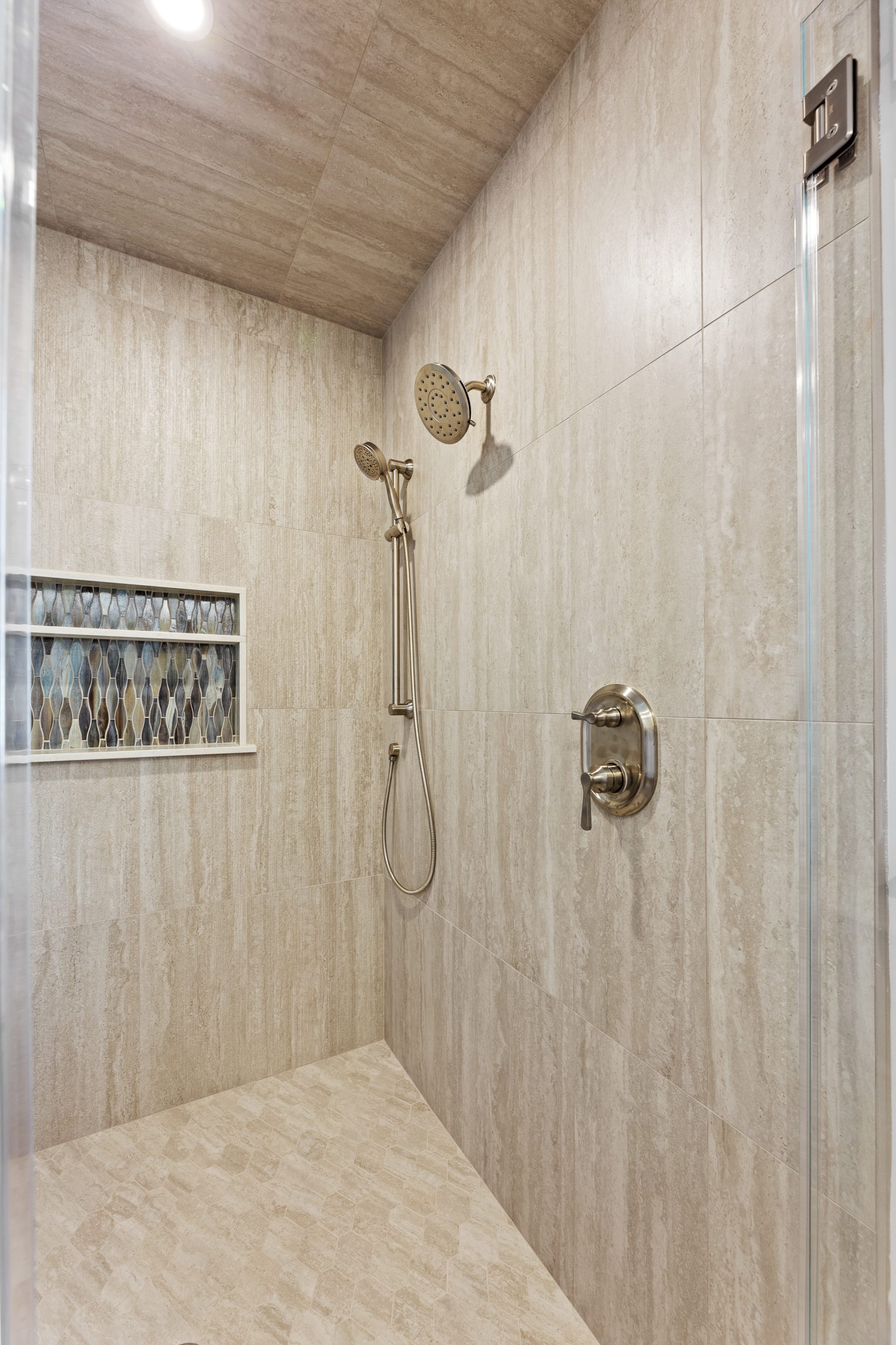 A modern walk-in shower with beige tile walls and floor, featuring a built-in niche with decorative glass, a rainfall showerhead, a handheld shower, and a metal control handle.