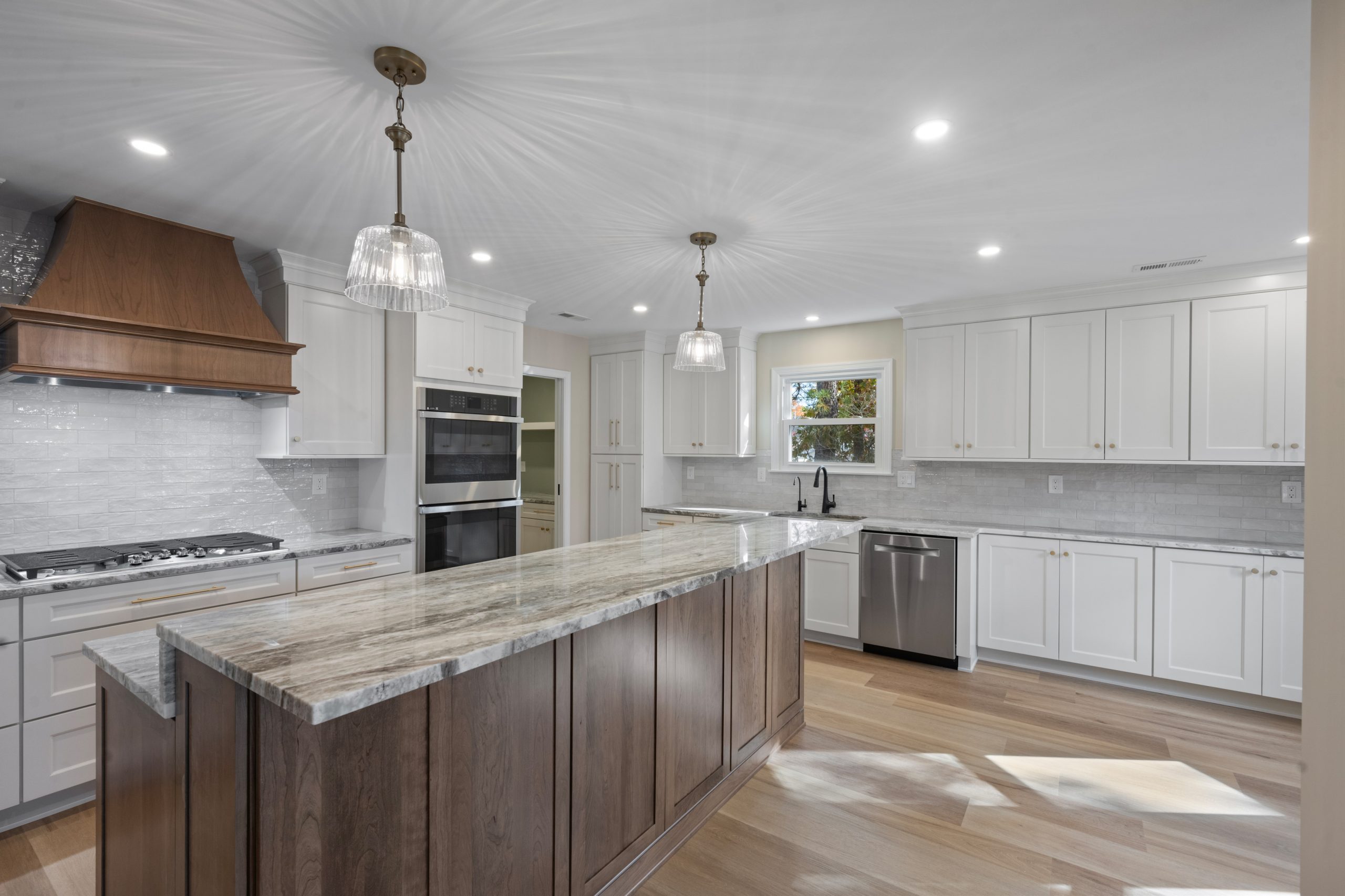 Modern kitchen with white cabinets, large granite island with wood base, stainless steel appliances, double ovens, and pendant lights. Sunlight streams through a window above the sink, highlighting the spacious layout.