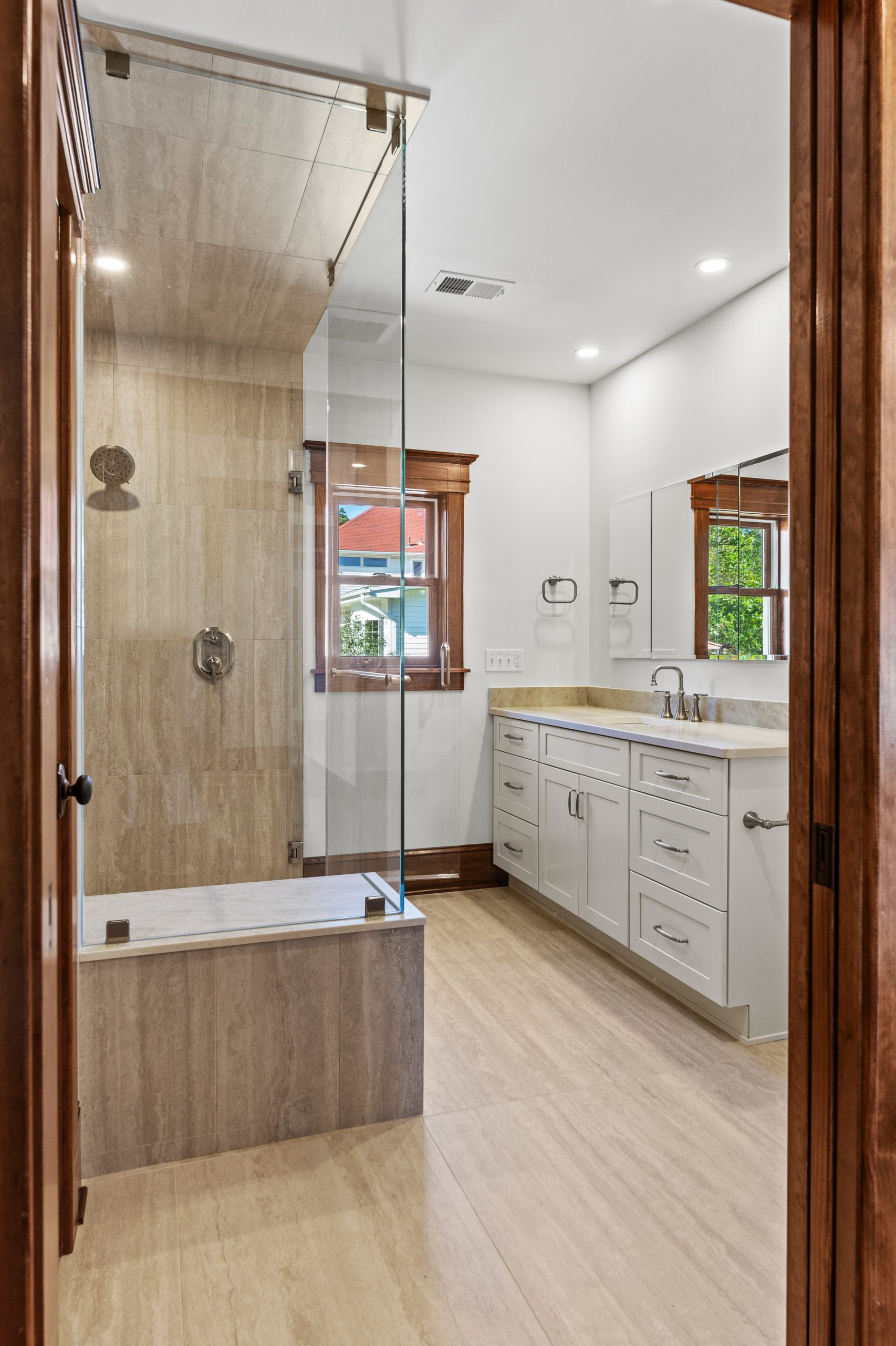 A modern bathroom with light wood cabinets, a large mirror, double sinks, and a spacious glass-enclosed shower featuring a built-in bench; wood trim accents the window and door frame.