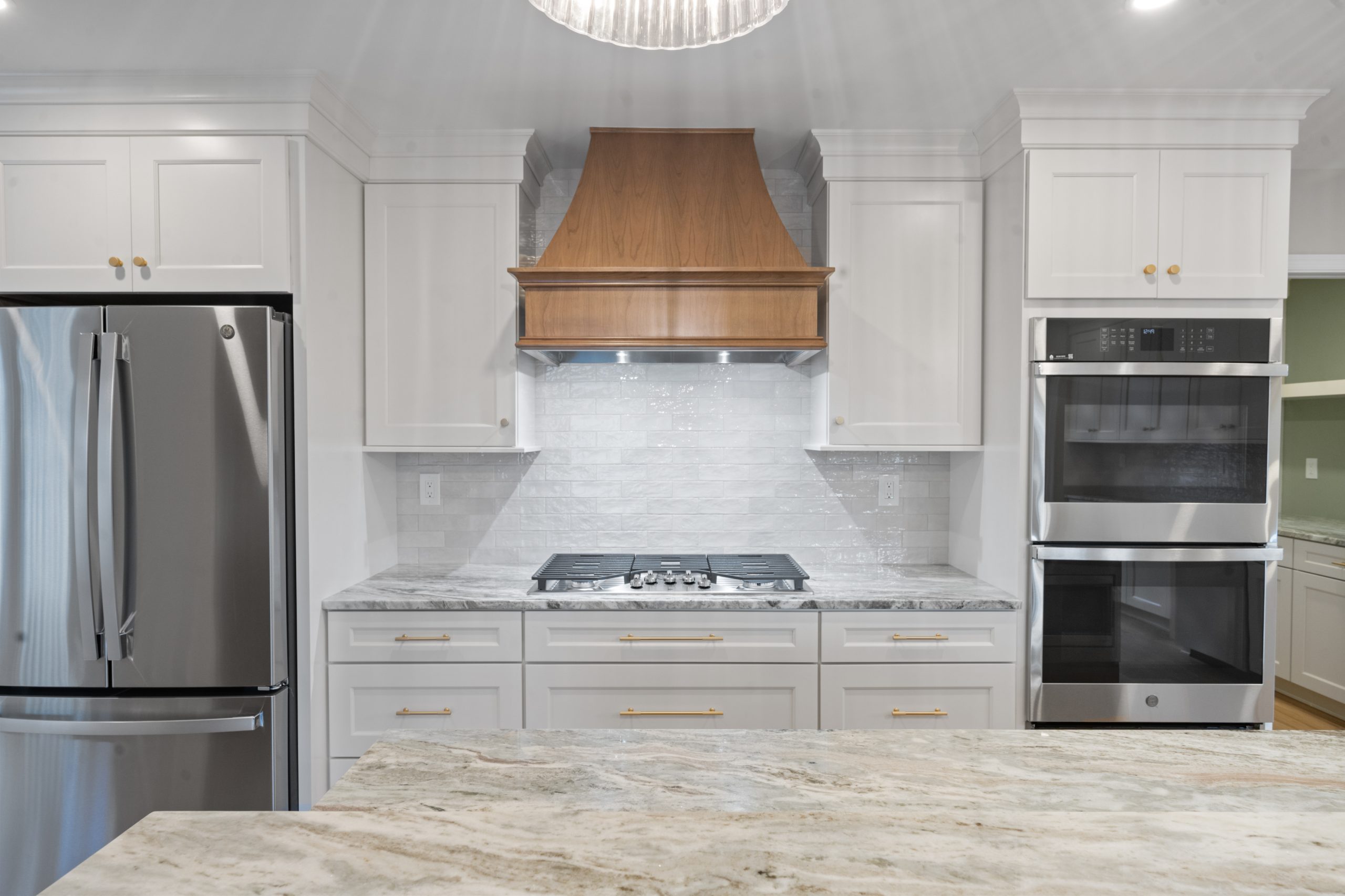 Modern kitchen with white cabinets, stainless steel refrigerator, double wall ovens, gas cooktop with a wooden range hood, gray marble countertops, and a tile backsplash under bright lighting.