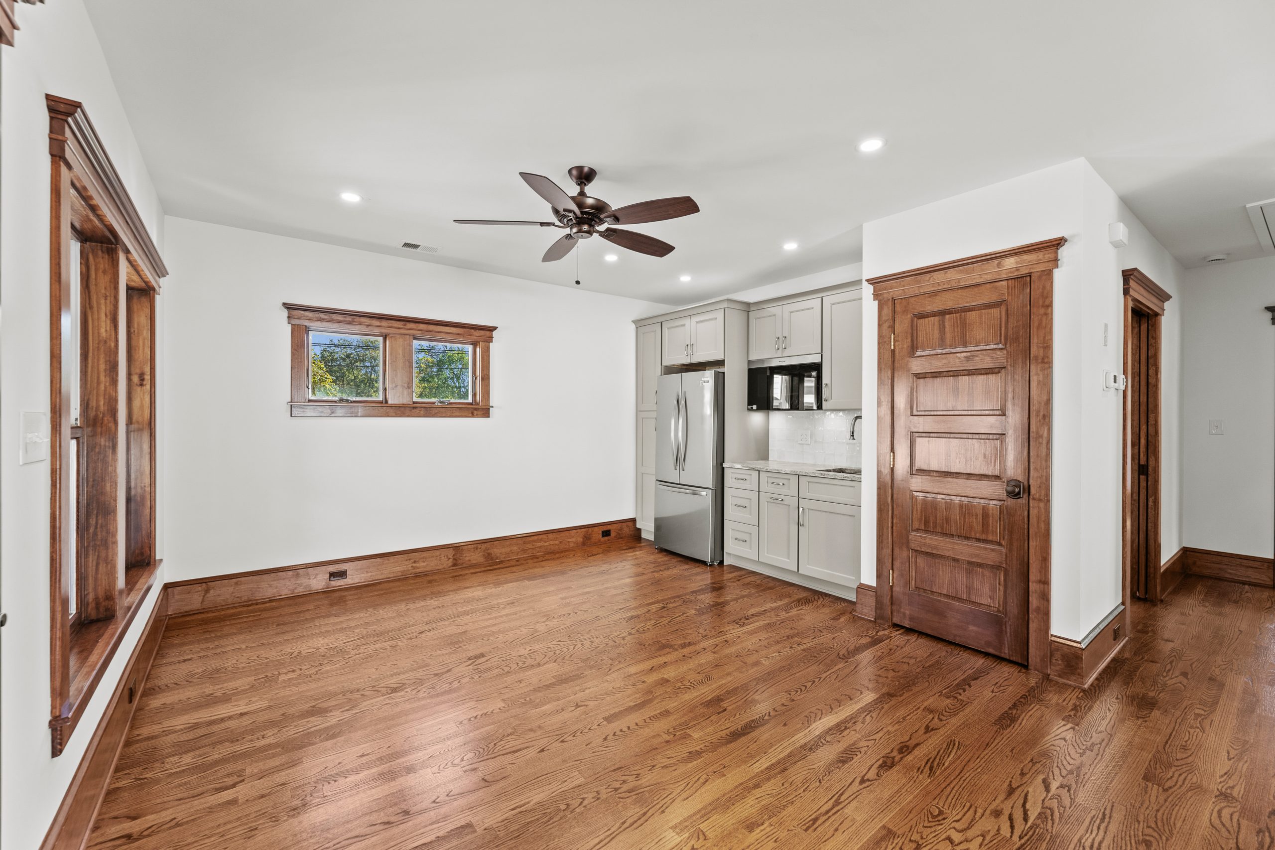 A bright, empty room with hardwood floors, a ceiling fan, and wood trim. The corner kitchen features white cabinets, a refrigerator, microwave, and stove, with two small windows above the sink.