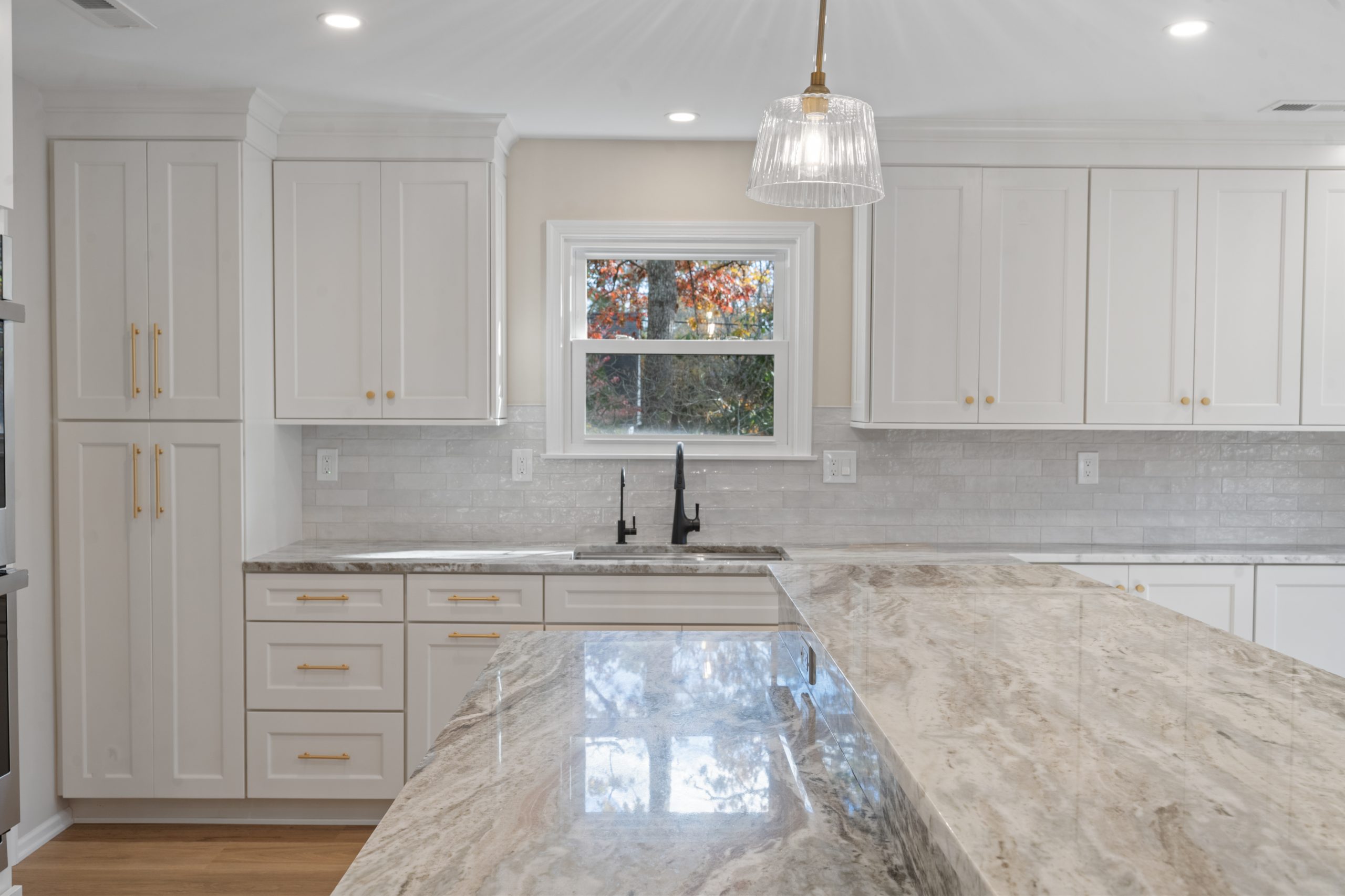 Bright, modern kitchen with white cabinets, gold hardware, marble countertops, a large island, and a window above a black faucet sink letting in natural light. A glass pendant light hangs from the ceiling.