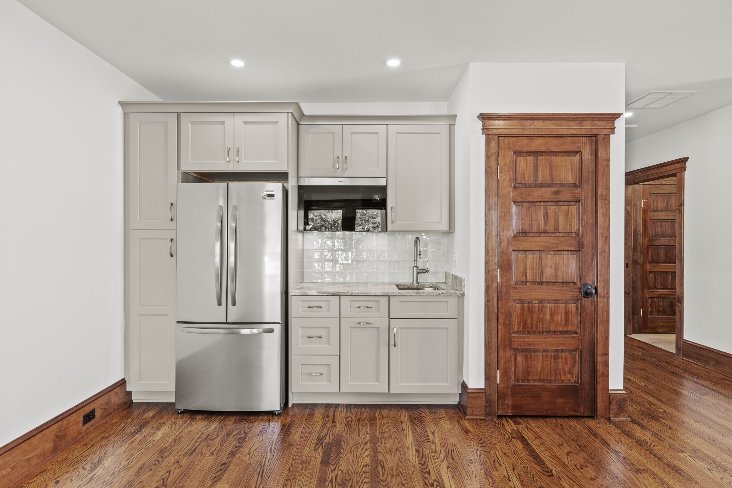 A compact kitchen with stainless steel French door refrigerator, light gray cabinets, white tile backsplash, and a small sink. The room has wooden floors and dark wood trim around the doors.