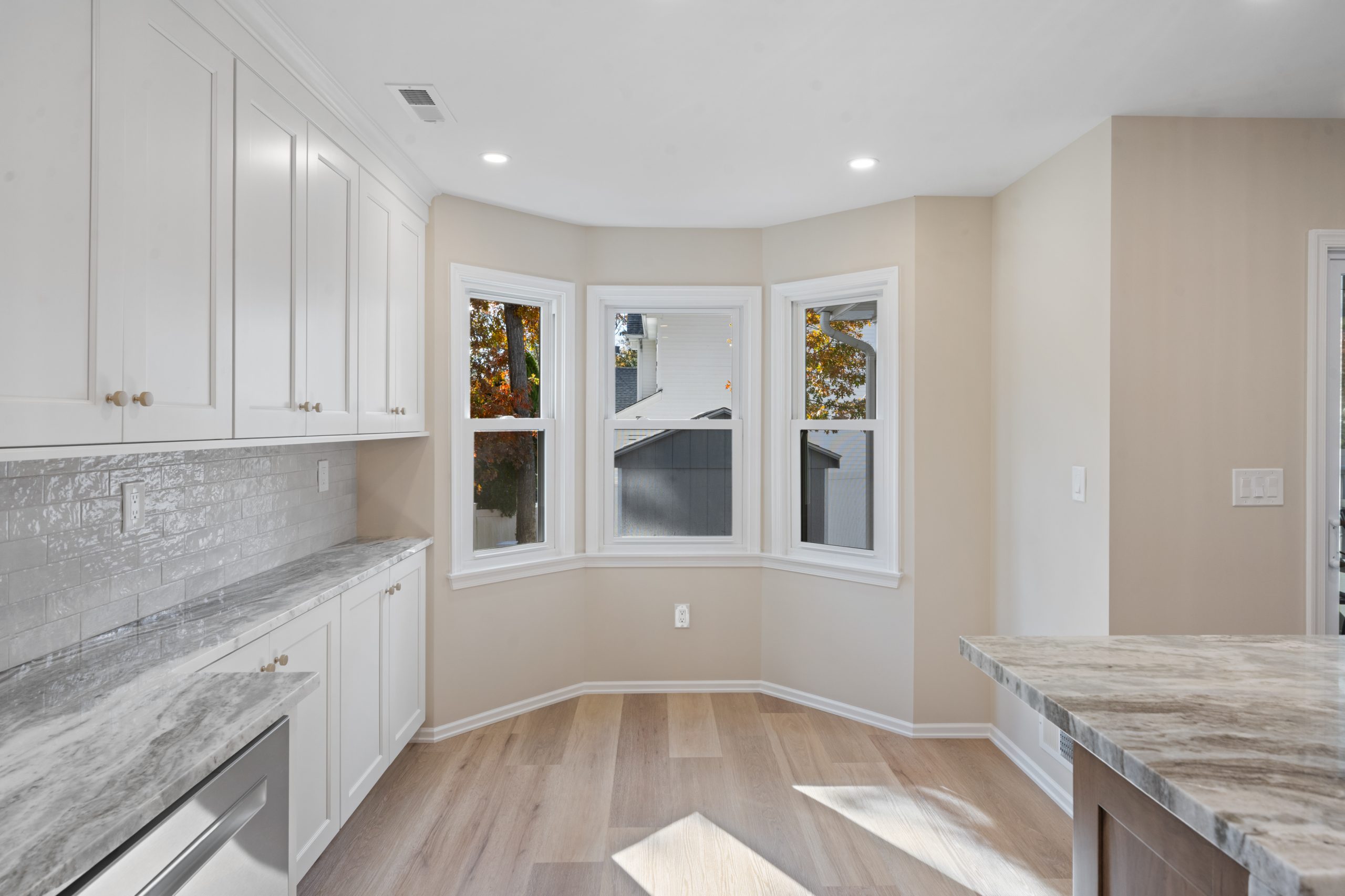 Modern kitchen with white cabinets, light wood flooring, gray stone countertops, and a bay window letting in natural light. Neutral walls and tiled backsplash create a bright, clean atmosphere.