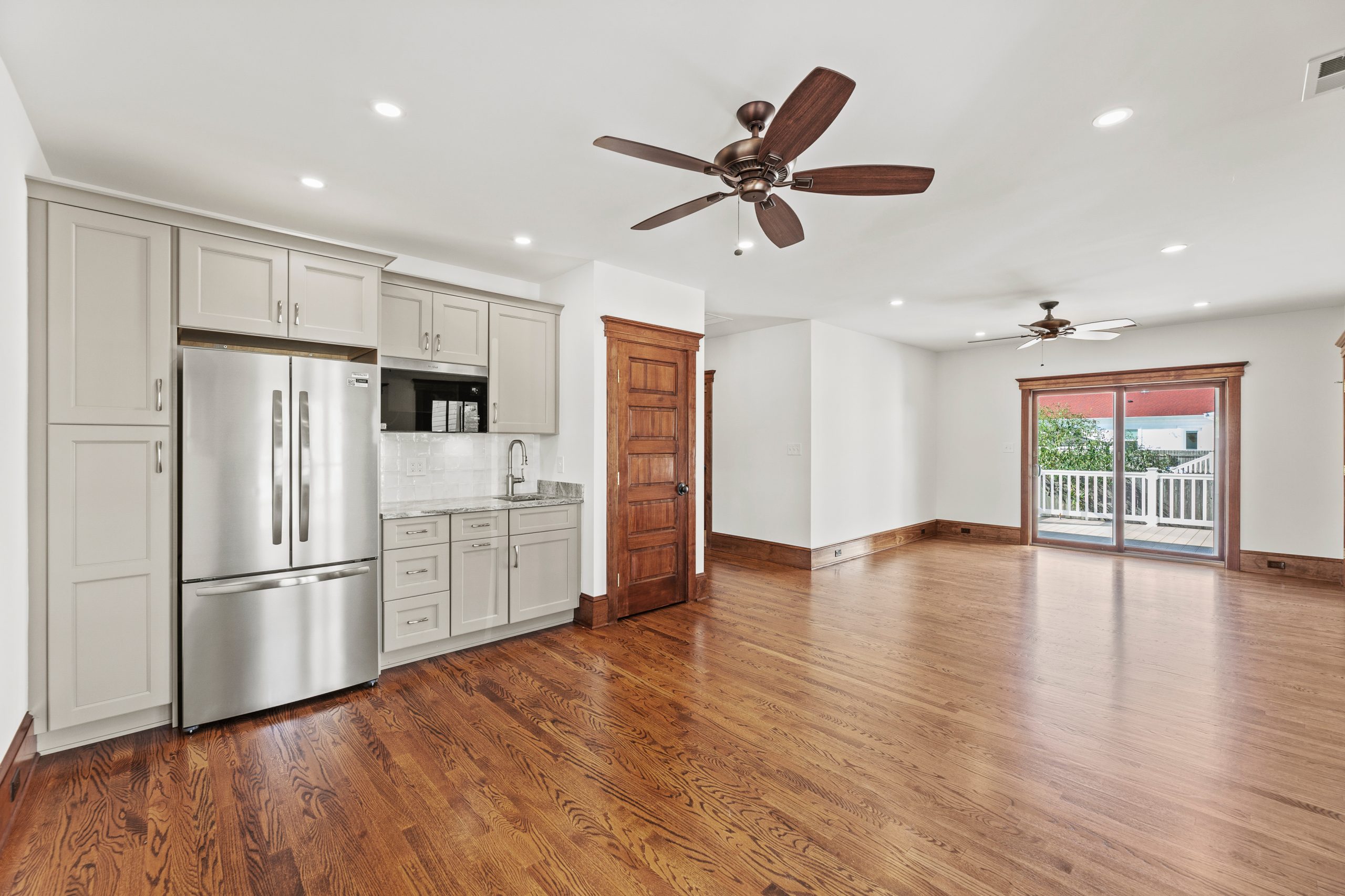 Spacious, bright room with hardwood floors, ceiling fans, a compact kitchen with stainless steel appliances, light gray cabinets, a wooden door, and large sliding glass doors leading to a balcony.