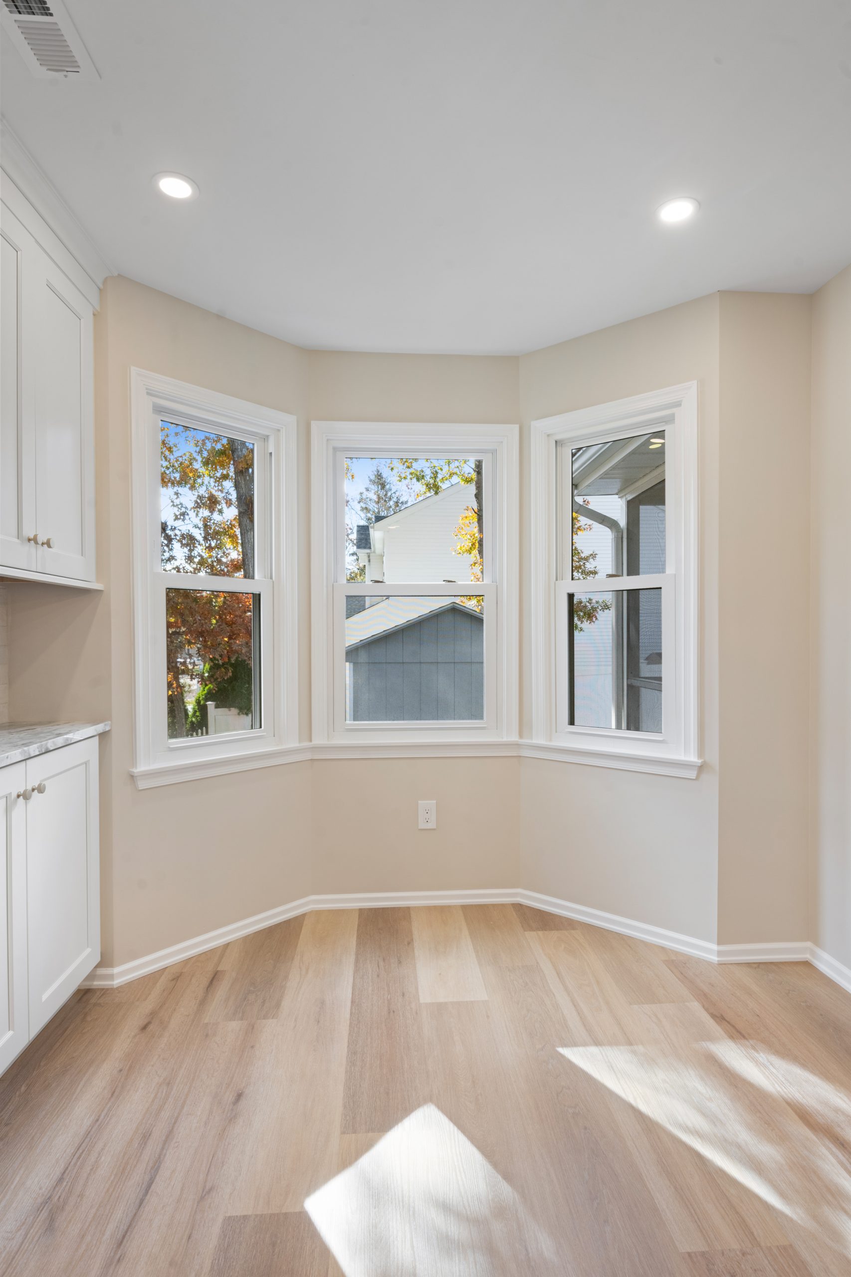 A bright room with light wood flooring, beige walls, and a bay window with three large panes overlooking trees and a neighboring house; white cabinetry is visible on the left.