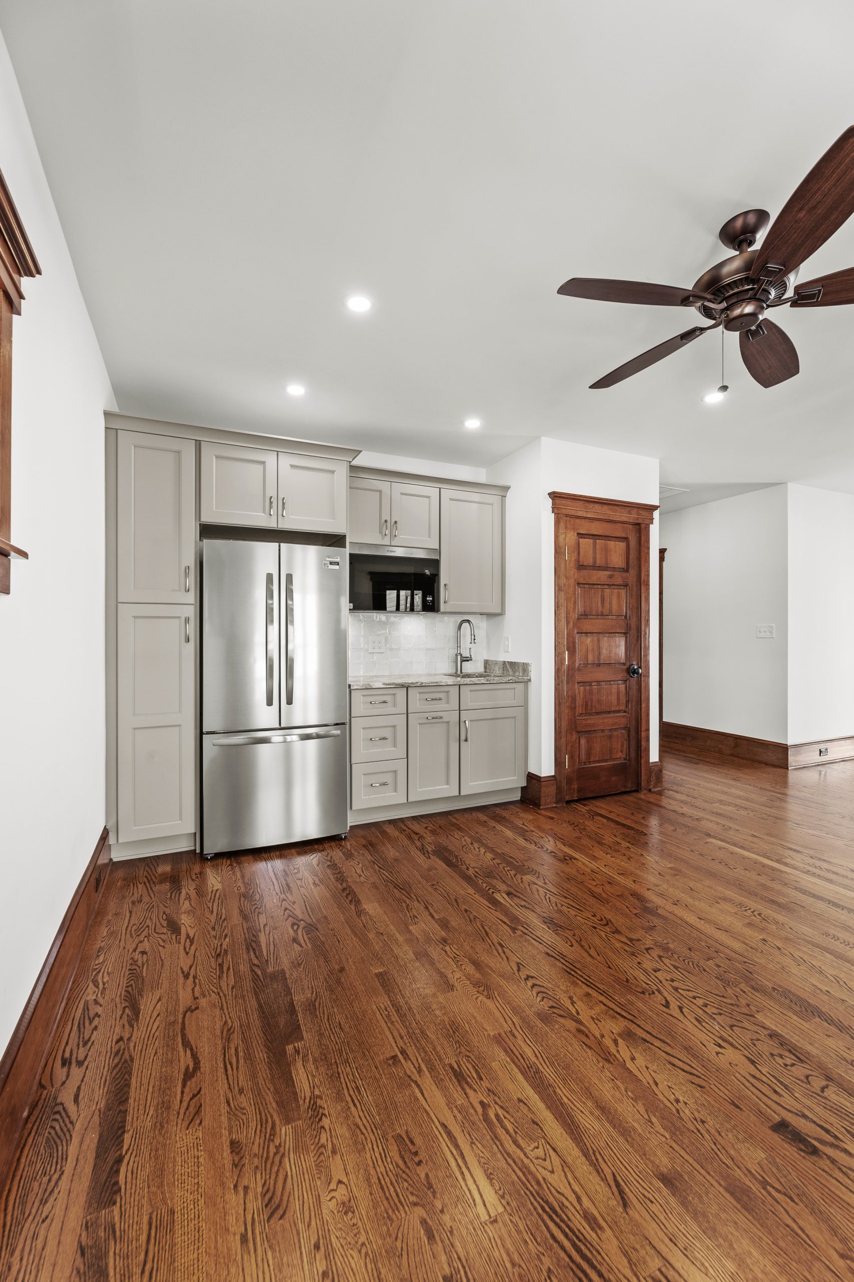 A kitchen area with light gray cabinets, stainless steel refrigerator, sink, white tile backsplash, and a wooden door. The room features hardwood floors and a ceiling fan with wooden blades.