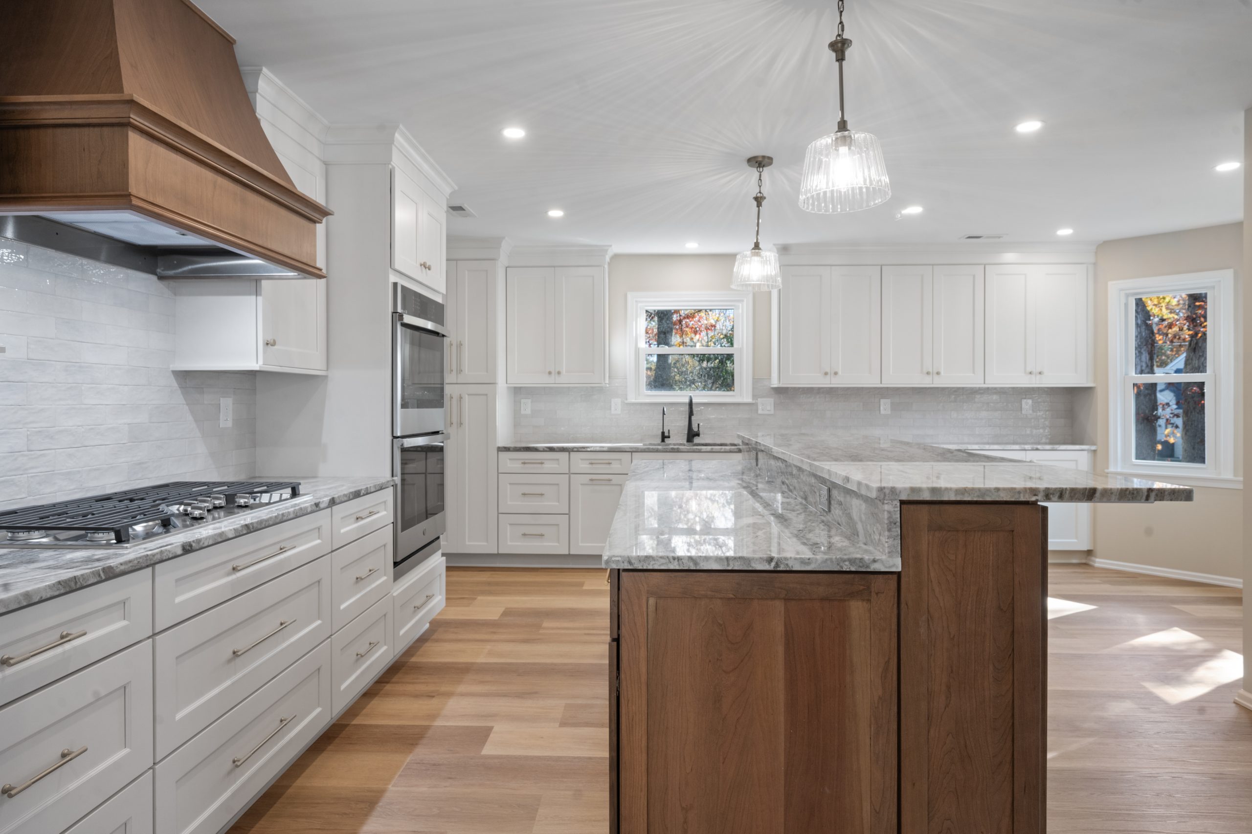 Modern kitchen with white cabinets, marble countertops, wood accents, a gas cooktop, double ovens, pendant lights, and light wood flooring. Two windows provide natural light and views of colorful autumn trees outside.