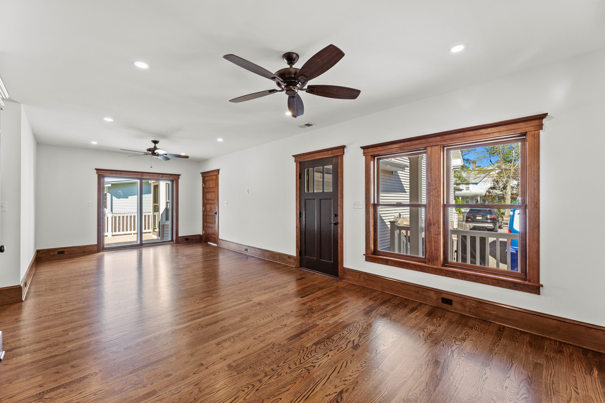 Bright, empty living room with wooden floors, large windows, two ceiling fans, a dark wooden front door, and a sliding glass door leading to a covered porch. White walls and wood trim create a warm, inviting atmosphere.