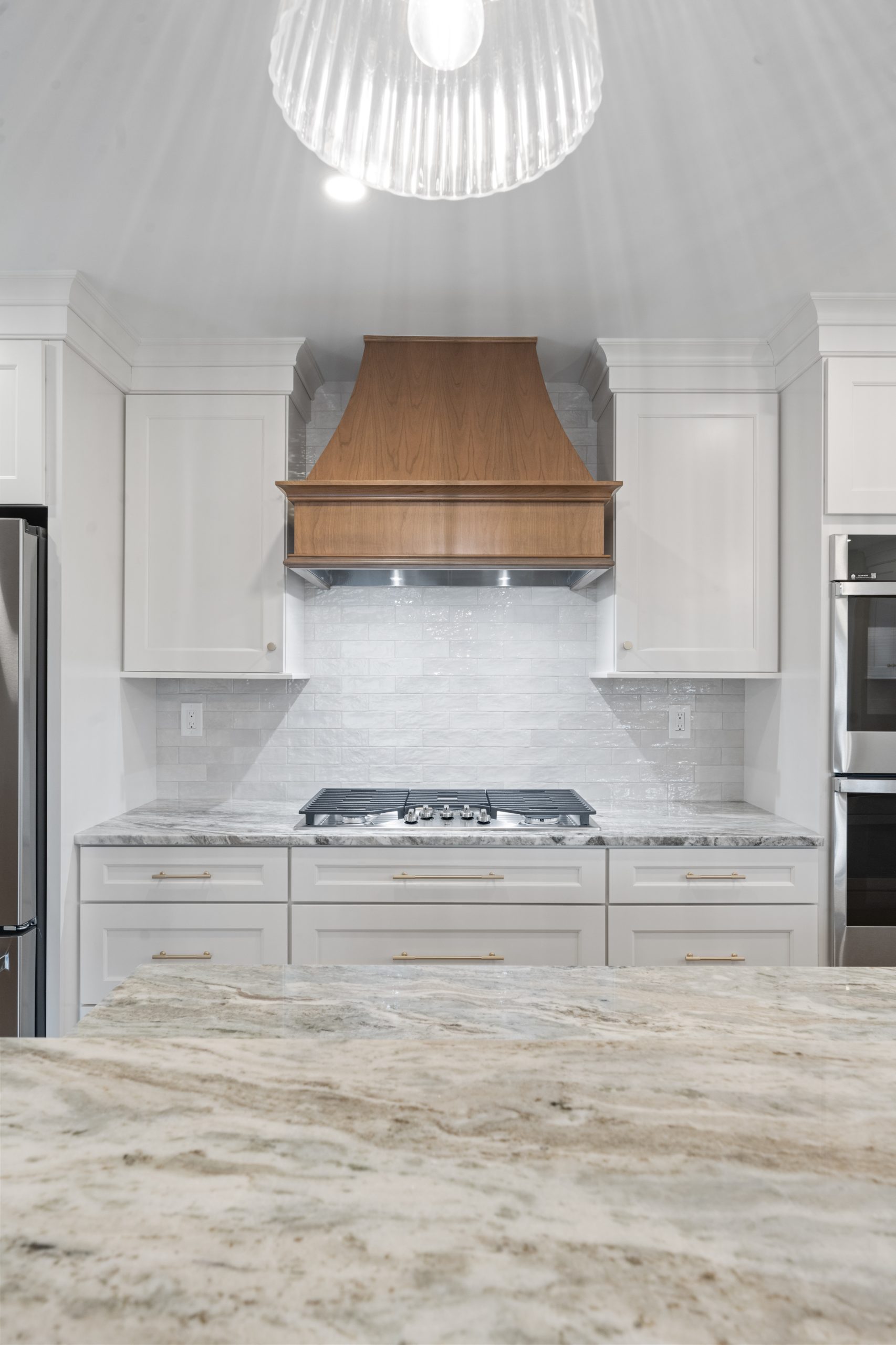 Modern kitchen with white cabinets, marble countertop, stainless steel appliances, and a wooden range hood above a stove. A pendant light hangs from the ceiling, casting light onto the scene.