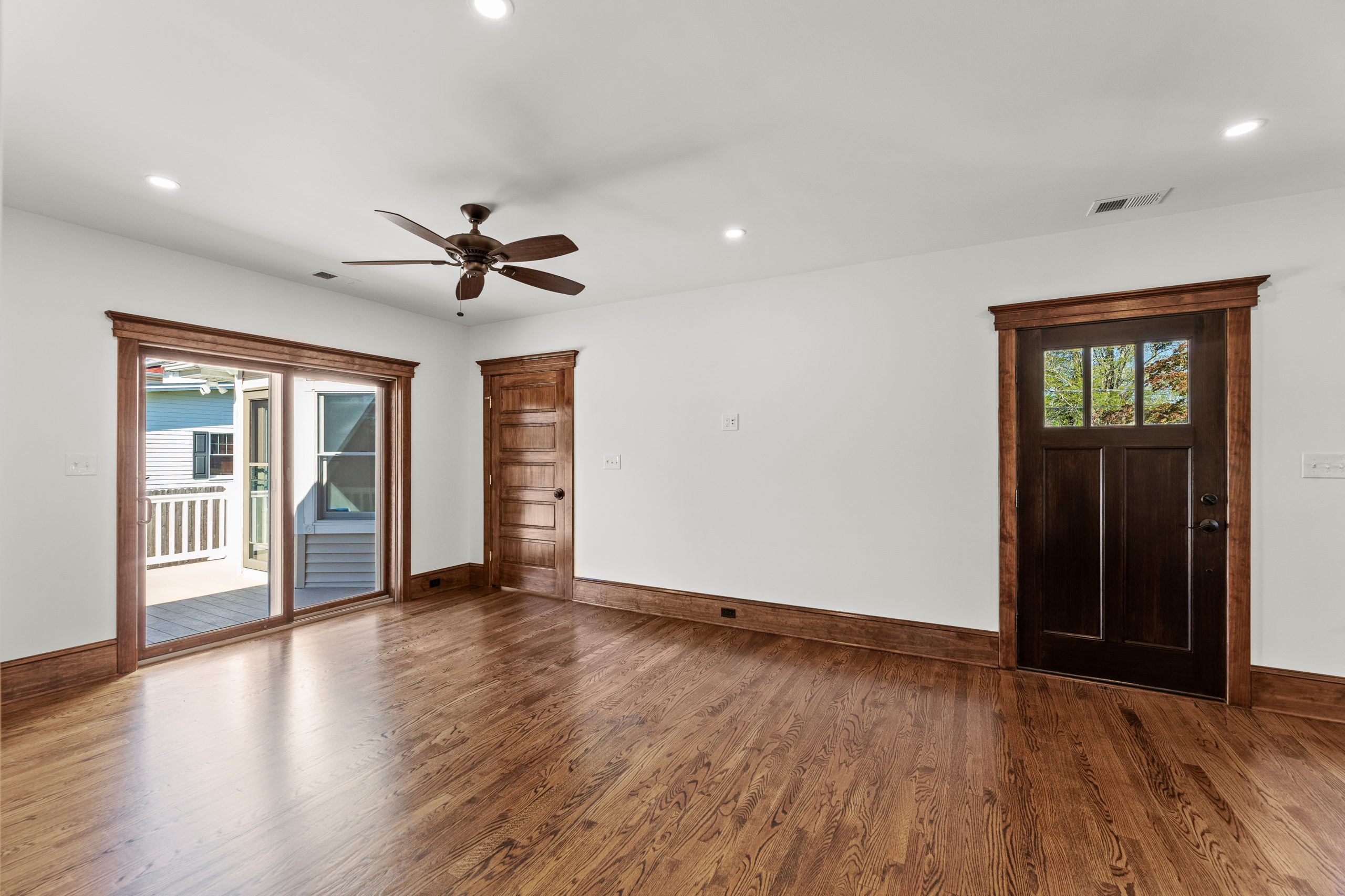 A bright, empty room with hardwood floors, white walls, and dark wood trim. There is a ceiling fan, a wood-framed glass door leading outside, a wooden door, and a windowed front door.